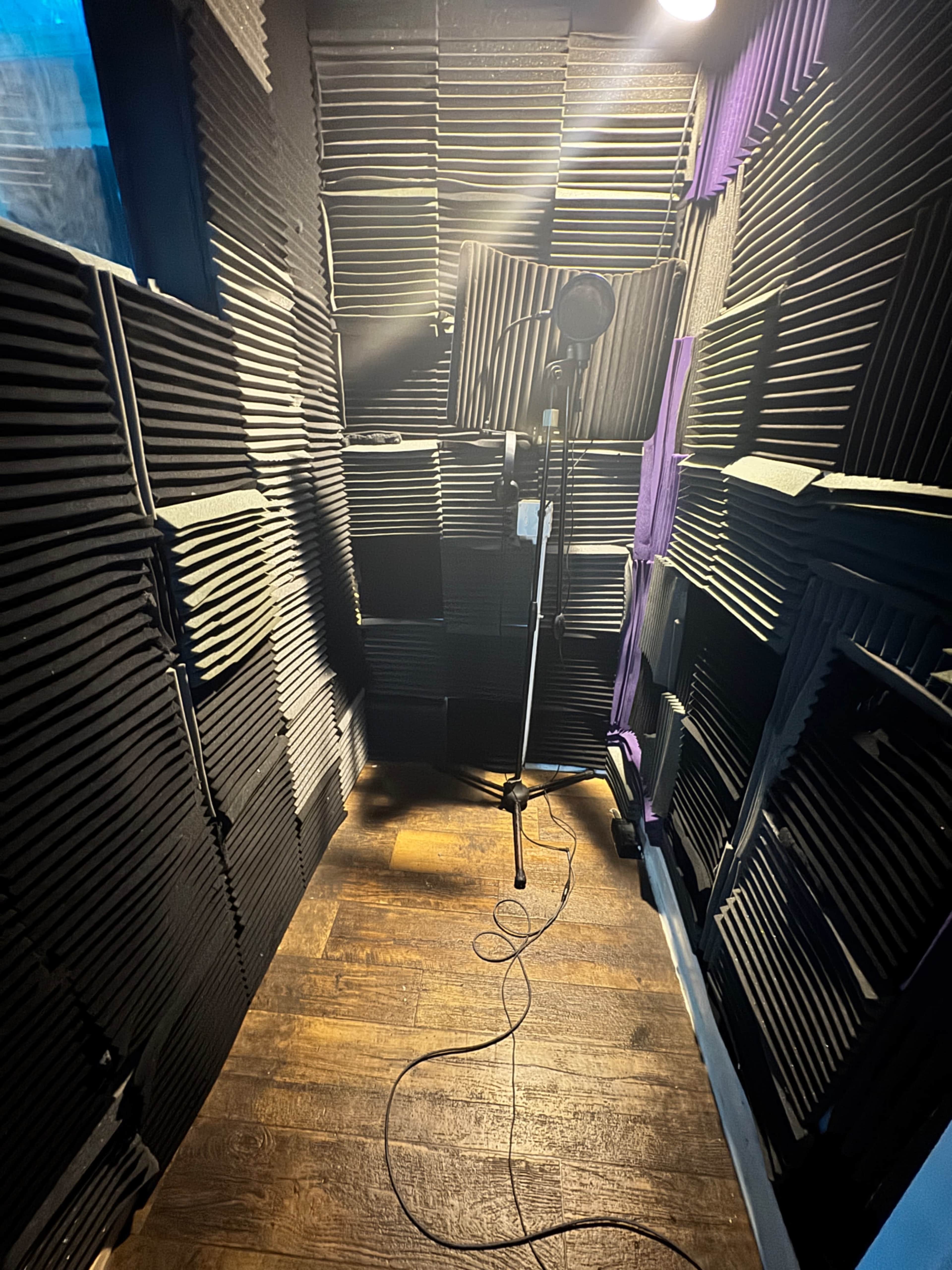 The image shows a soundproof recording booth lined with acoustic panels and a microphone on a stand, with a cable running along the floor.