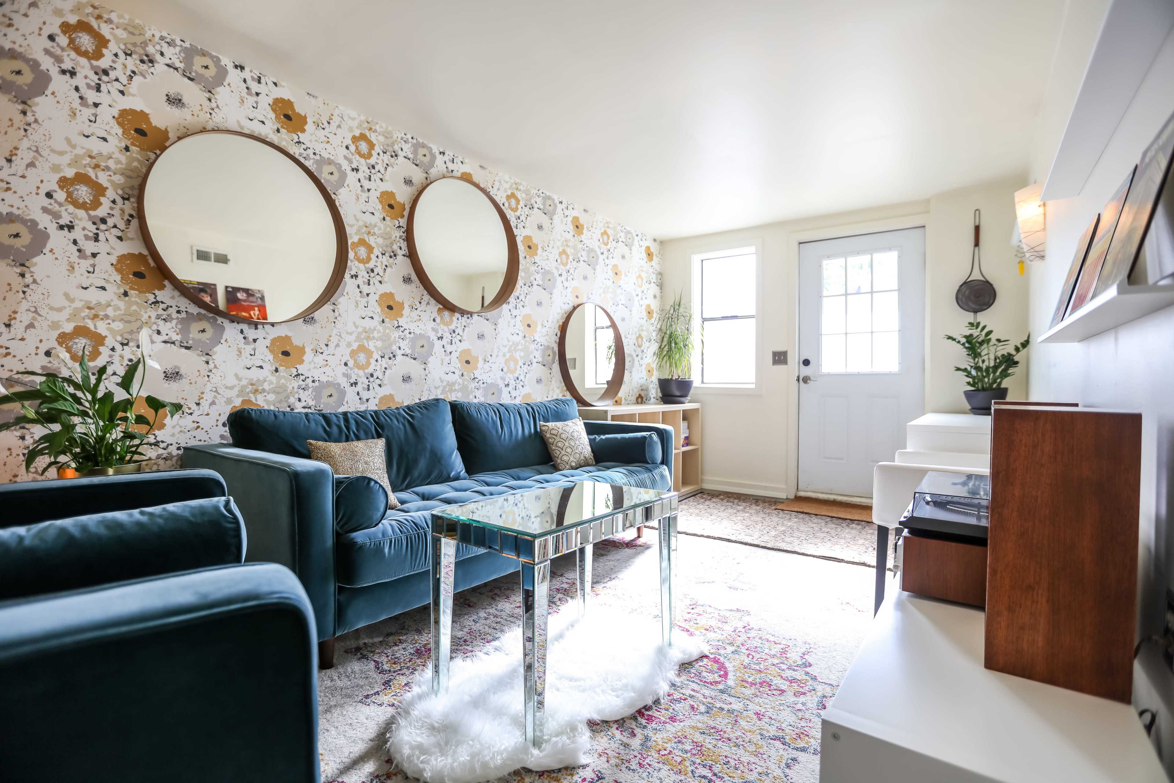 A well-decorated living room featuring a teal sofa, circular mirrors on the floral wallpaper, and a glass coffee table near a door with a small window.