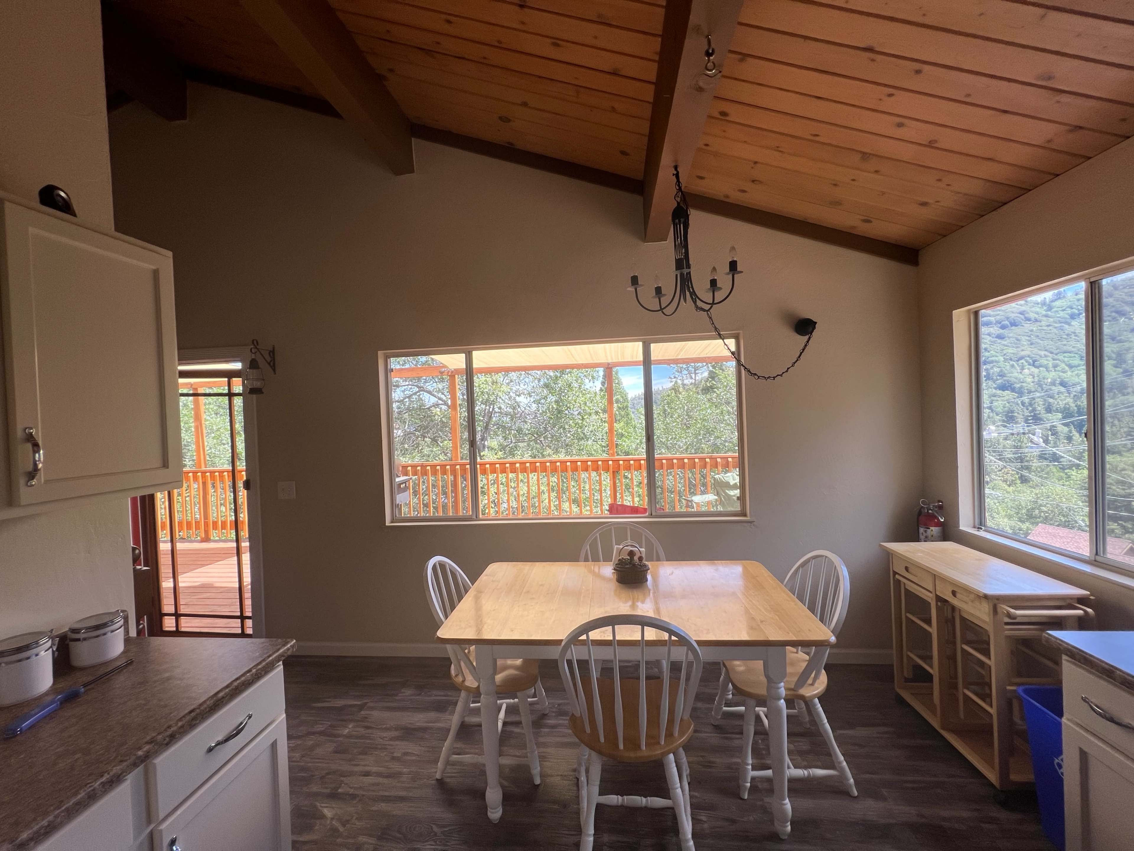 A spacious kitchen features a wooden table surrounded by chairs, with large windows offering a view of a deck and trees outside.