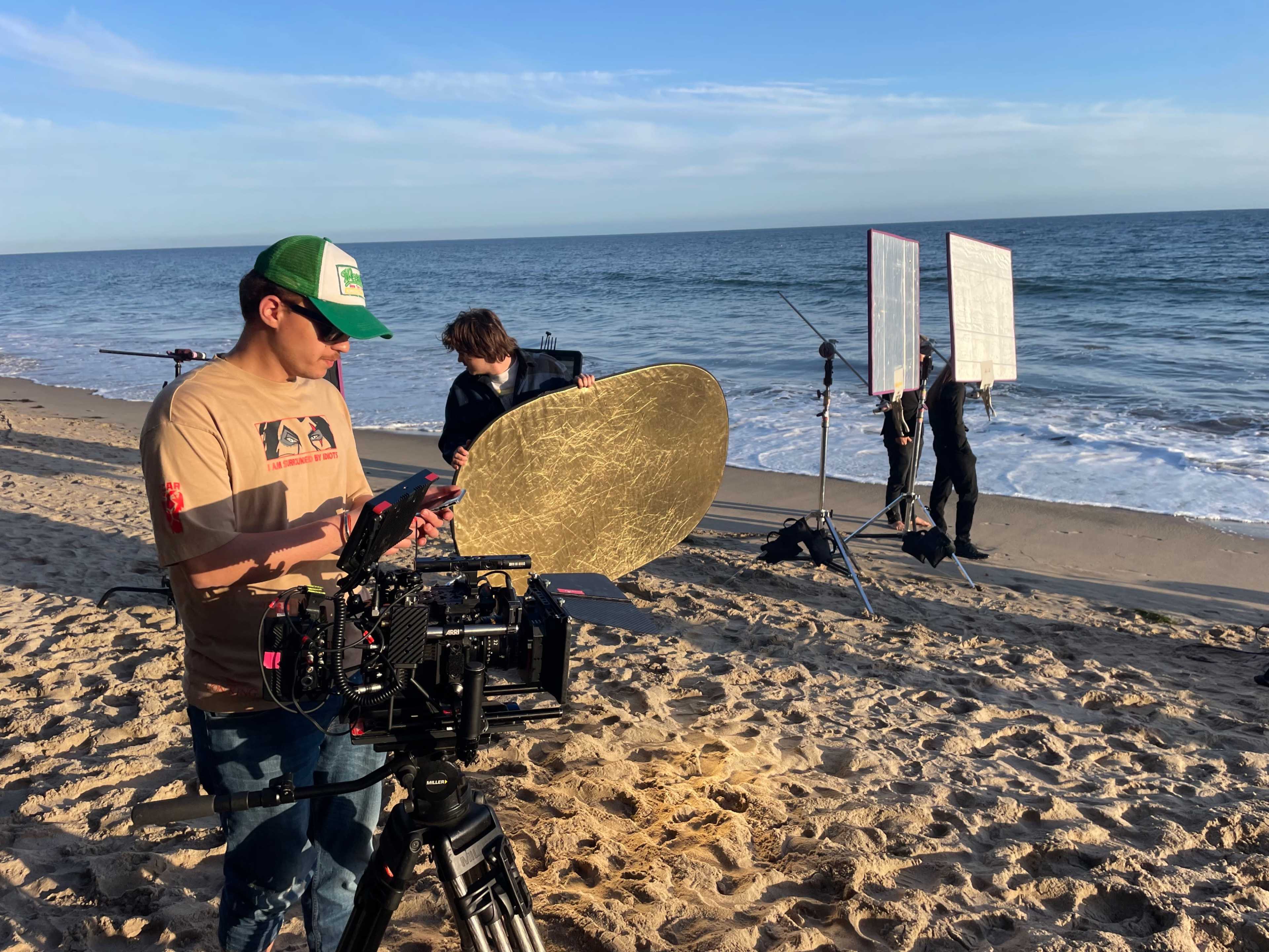 A film crew sets up equipment on the beach near the ocean, with a camera on a tripod and reflectors positioned for lighting.