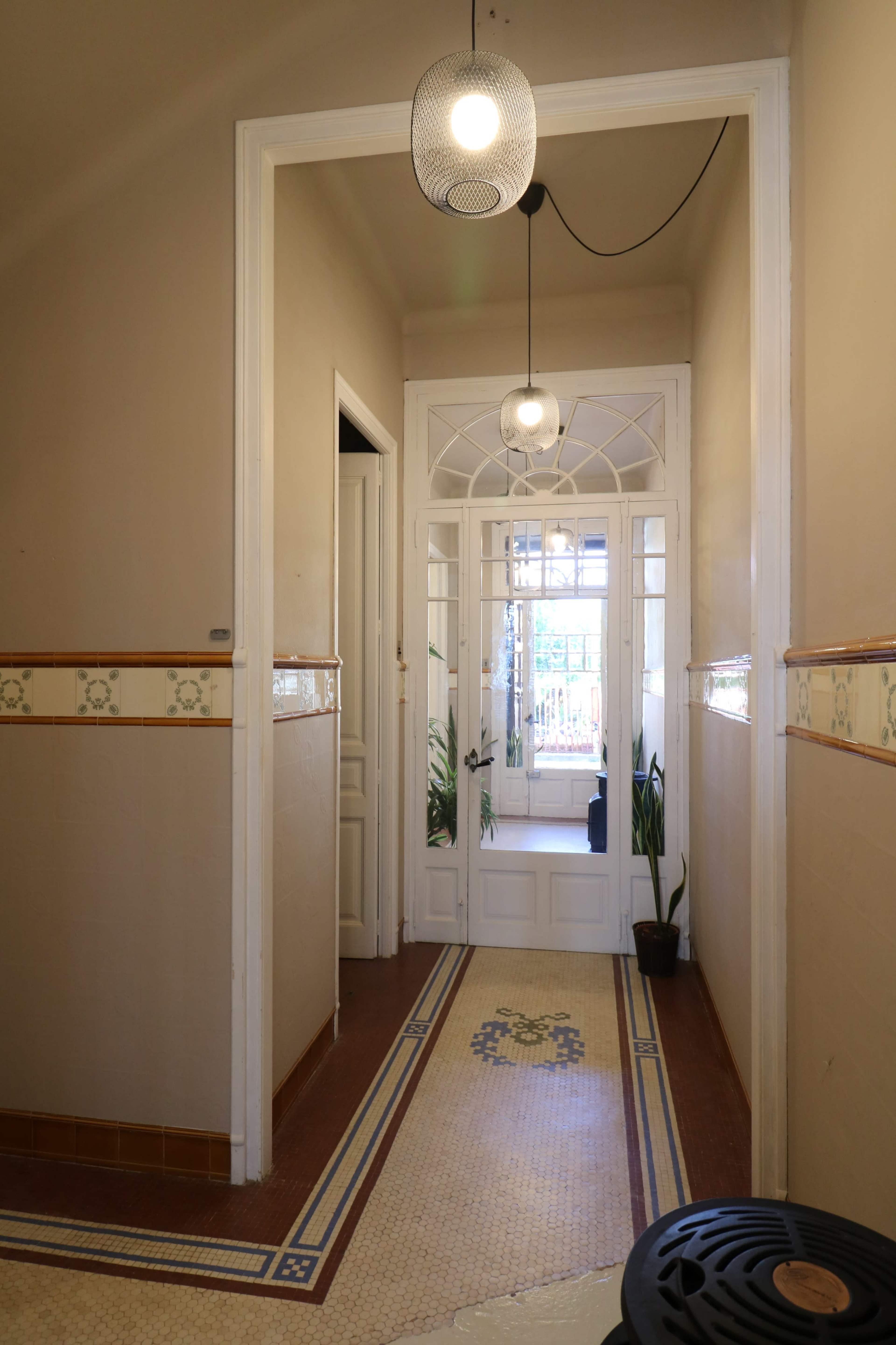 A hallway with tiled flooring, decorative wall tiles, and a glass door leading to a bright area with potted plants.