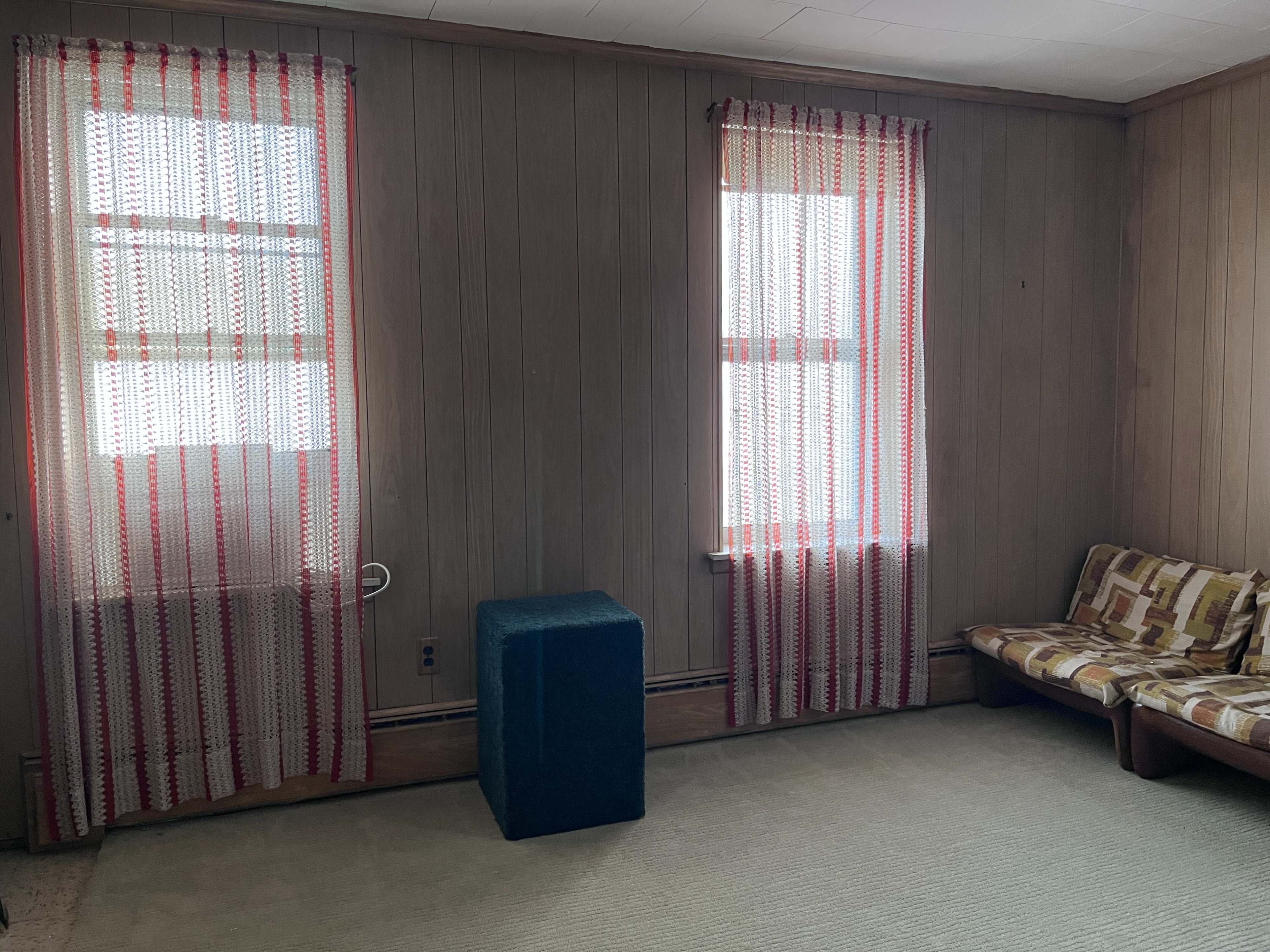 The room has beige paneling, two windows with striped curtains, a blue cube-shaped object, and a small couch with a patterned design.