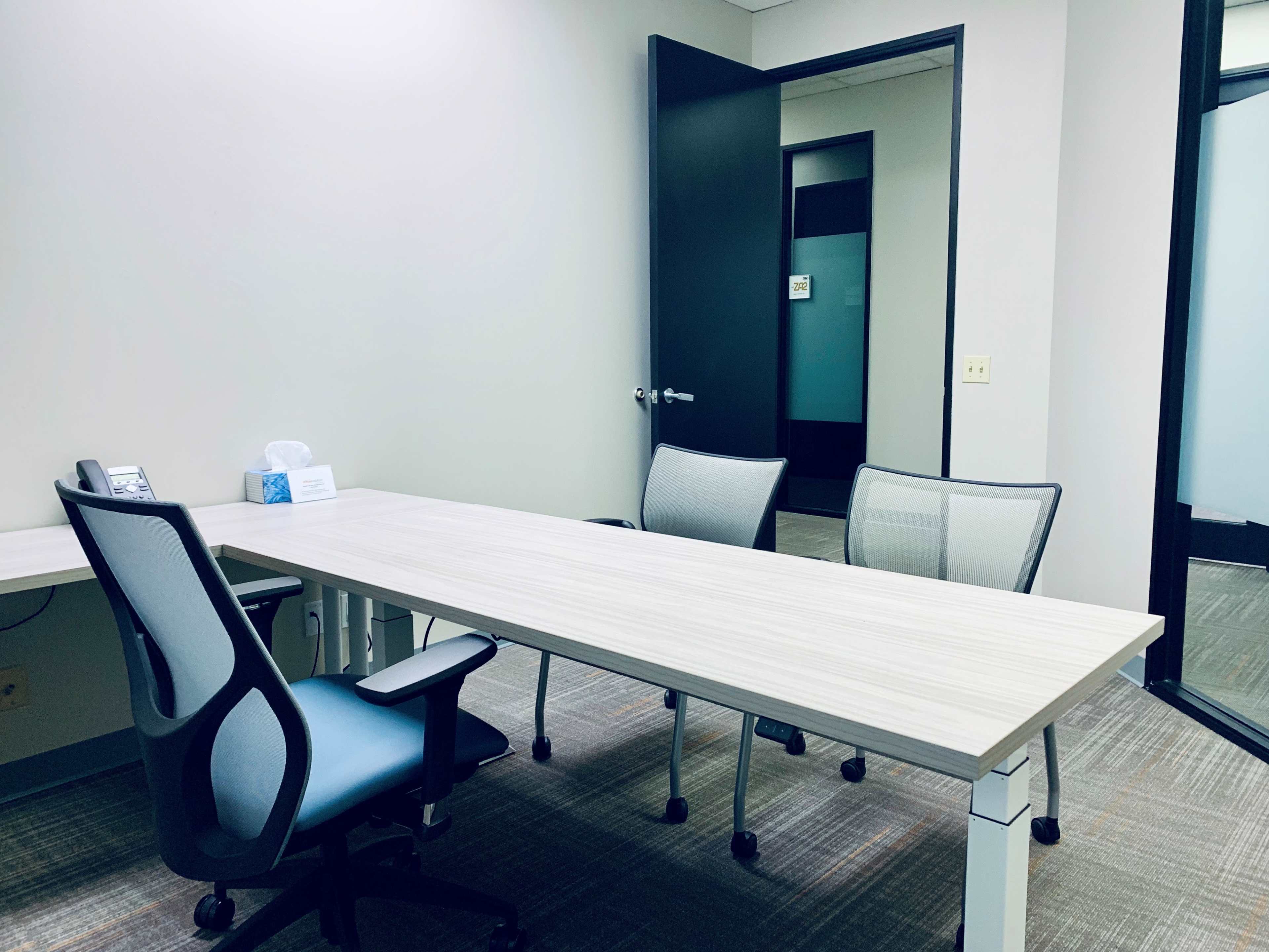 The image shows a small office with a long wooden table, three chairs, a phone, and an open door leading to another room.
