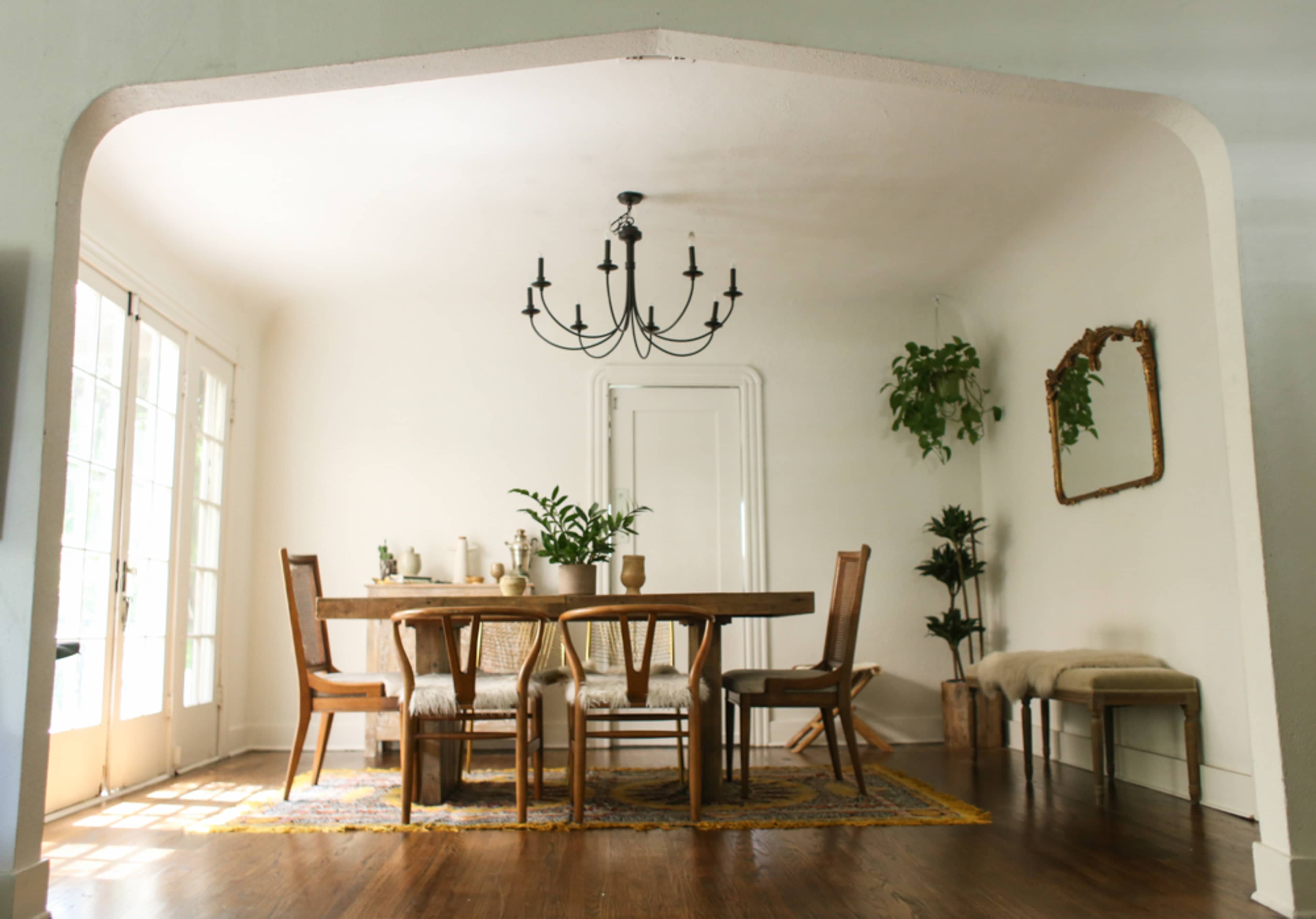 A spacious dining room features a large wooden table surrounded by chairs, with a chandelier overhead and plants along the walls.