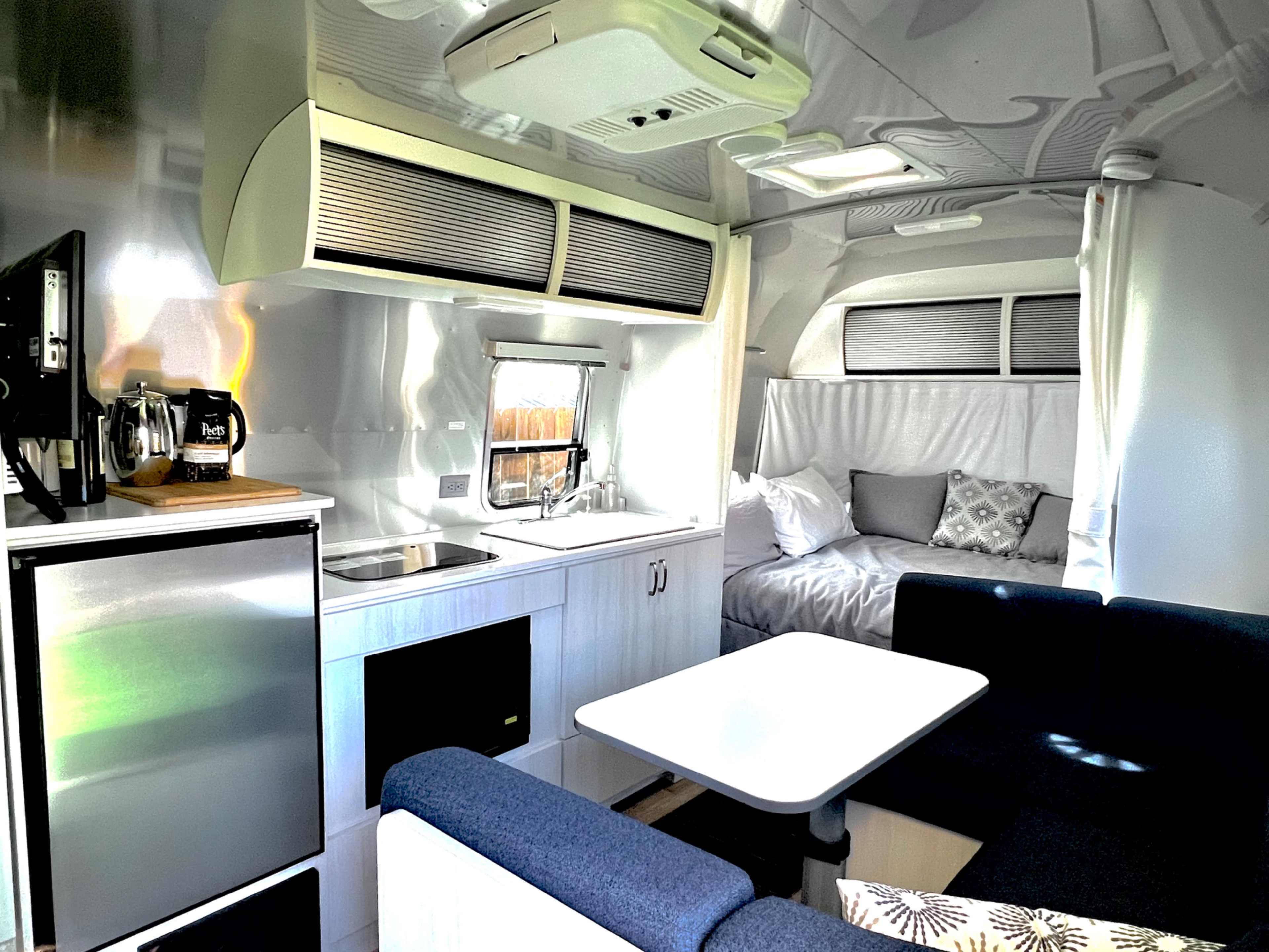 The image shows the interior of a modern camper trailer featuring a kitchenette, a dining area, and a sleeping space.