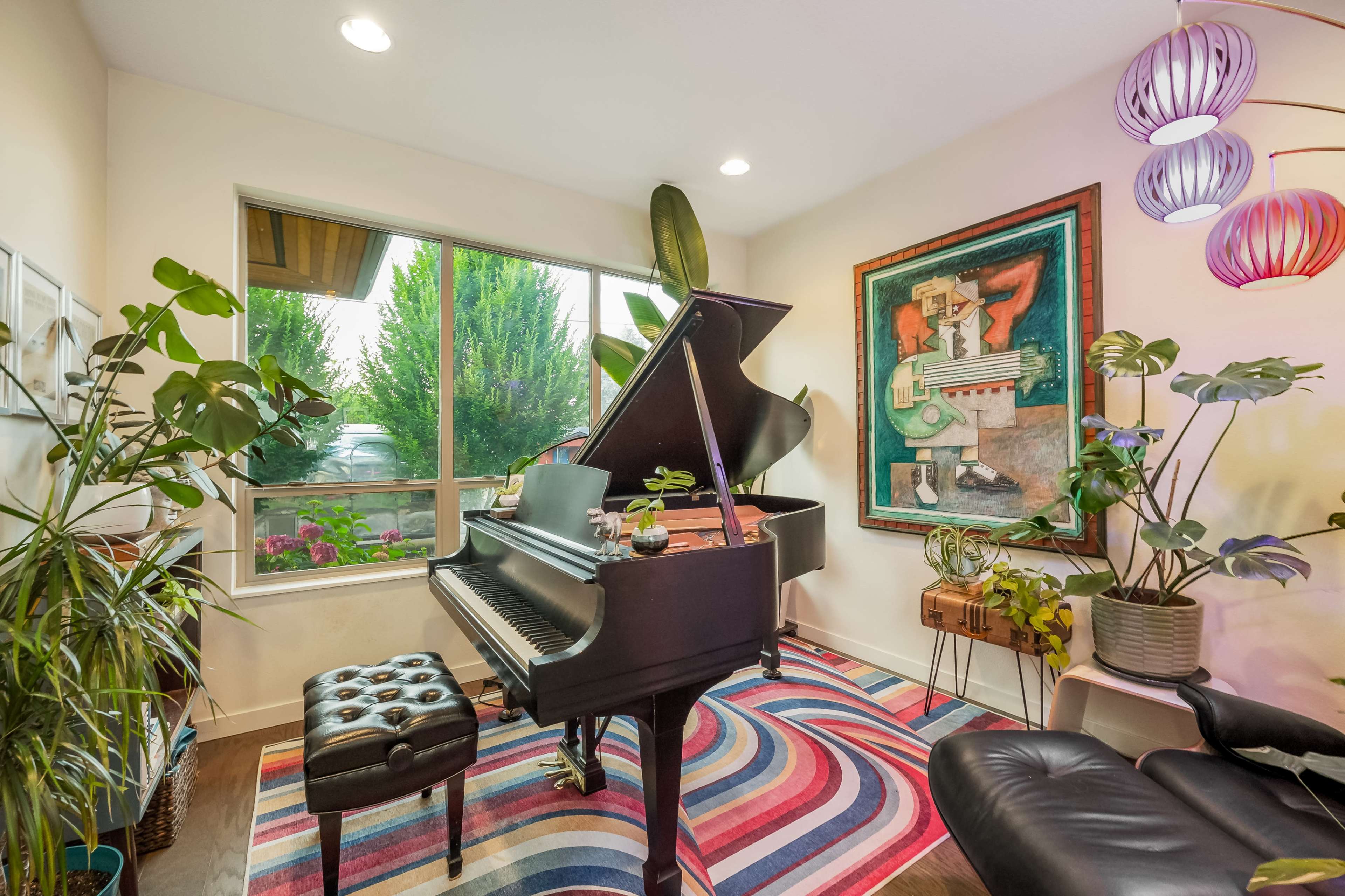 A grand piano sits next to a large window adorned with plants and vibrant artwork in a cozy room.