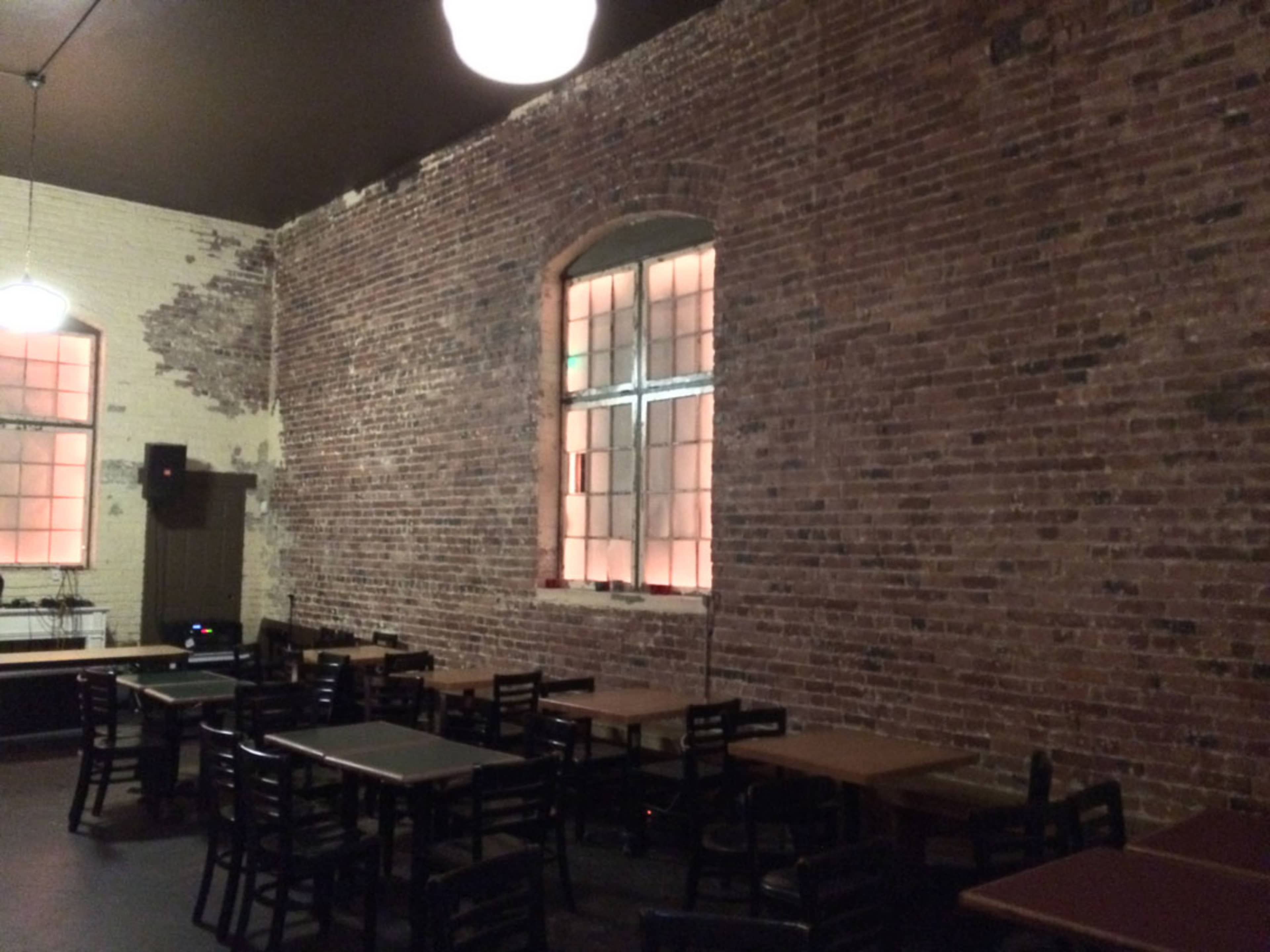 An interior space with exposed brick walls, empty tables, and a large window illuminated from the outside.