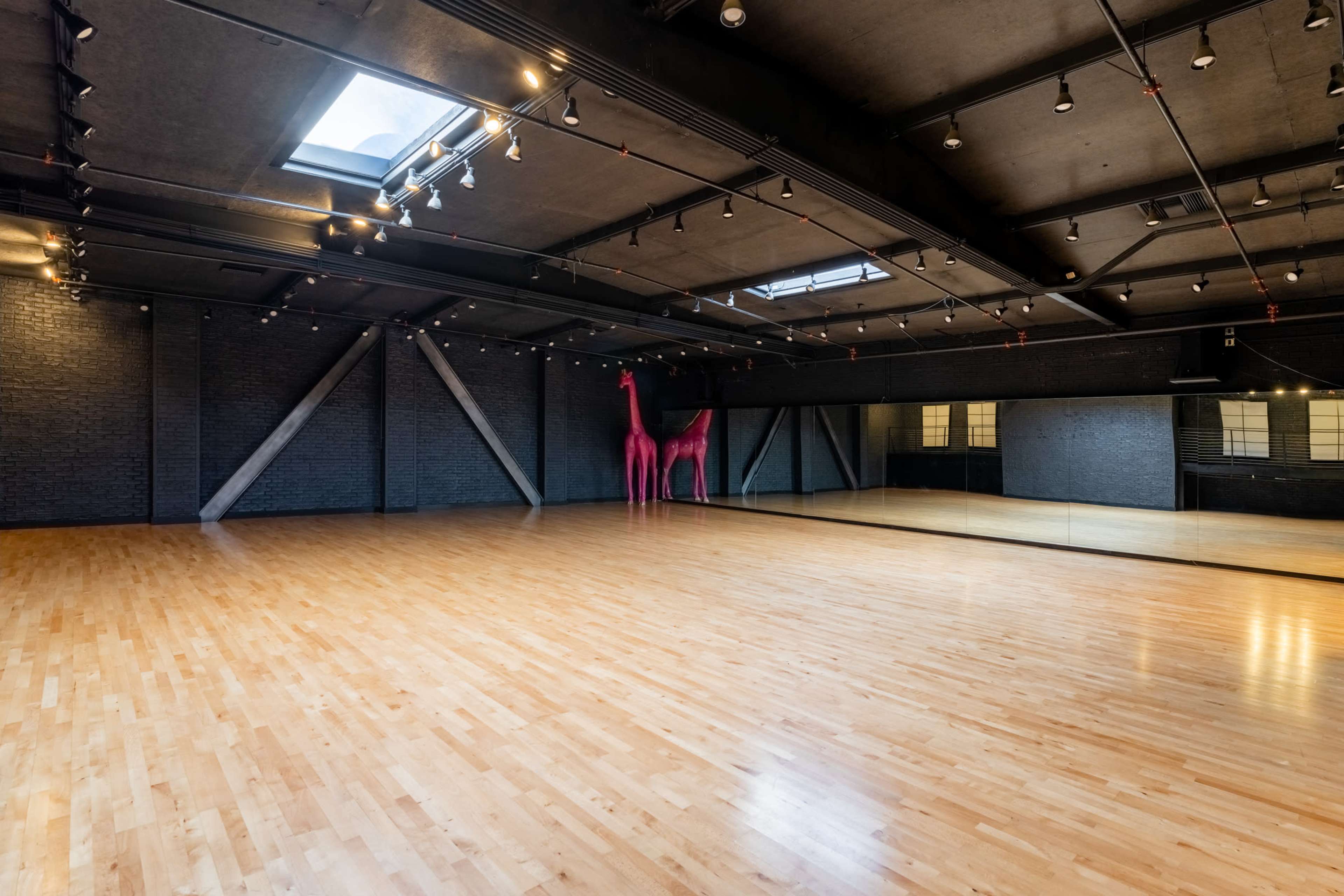 A spacious empty dance studio with a wooden floor, large mirrors, and a pink giraffe sculpture against a dark brick wall.