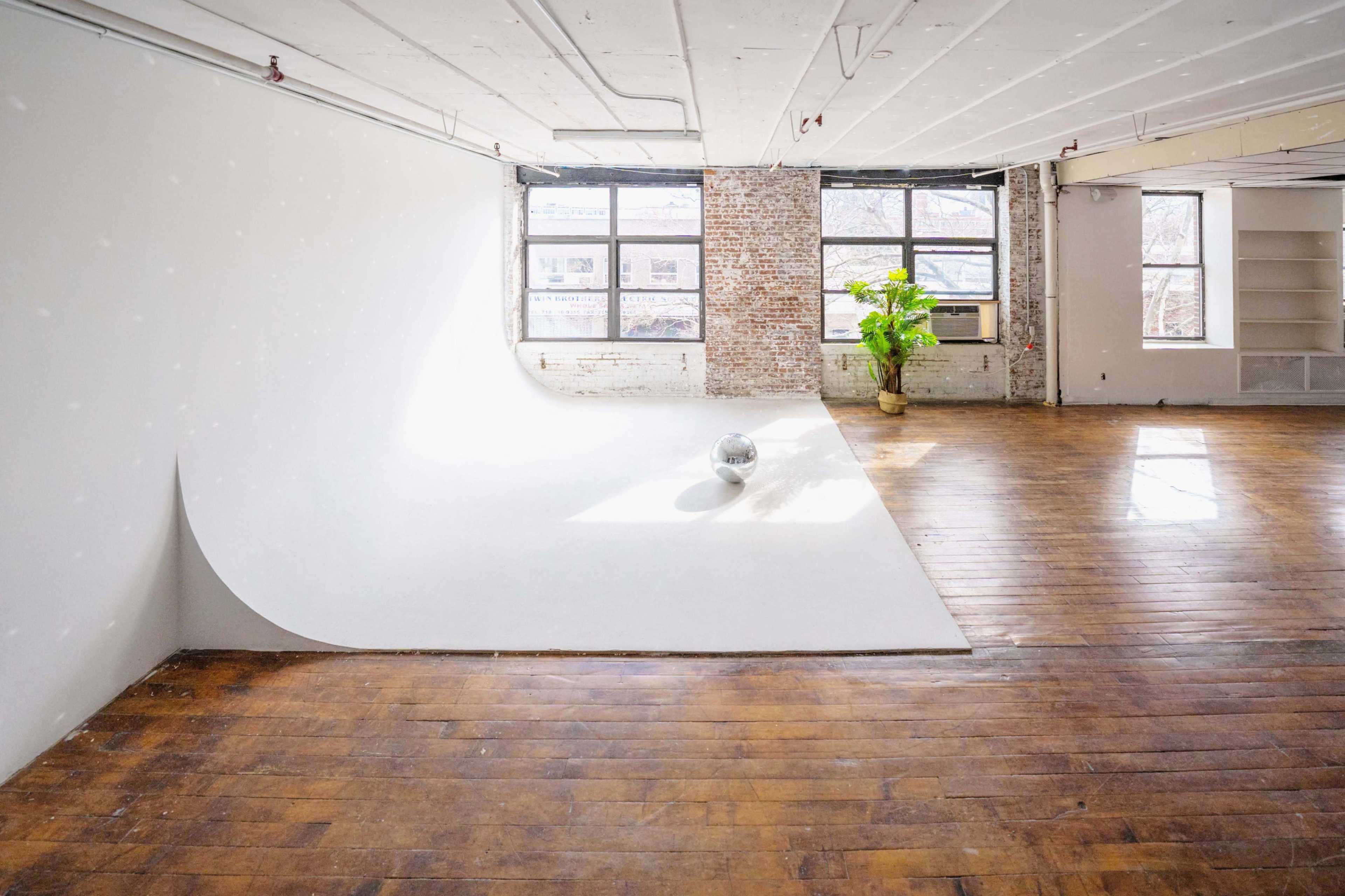A spacious, minimalist room with large windows, a smooth white wall curving into the floor, a solitary reflective sphere, and a potted plant near the windows.