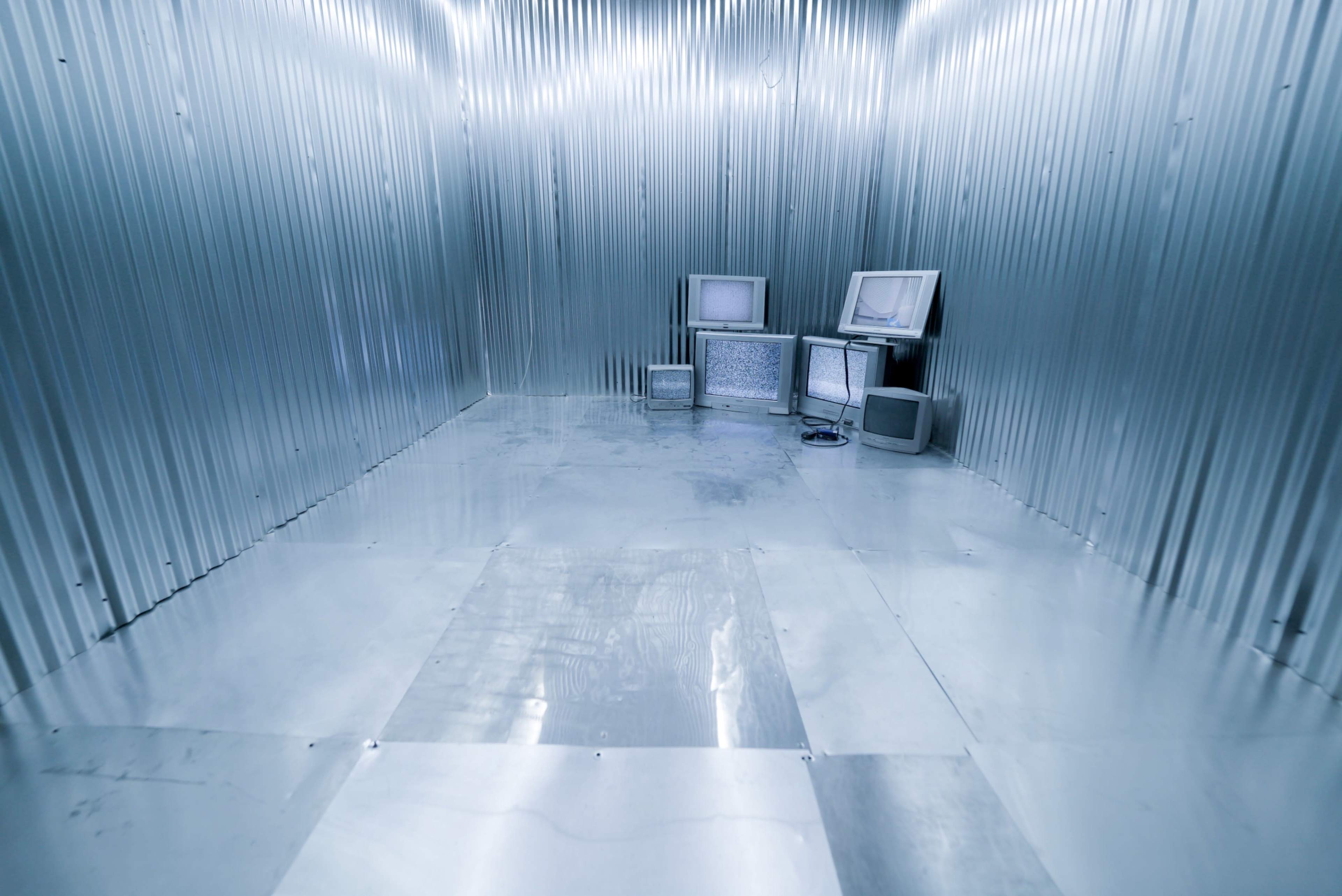 The image depicts a metallic room with corrugated walls, featuring several old televisions arranged in one corner.