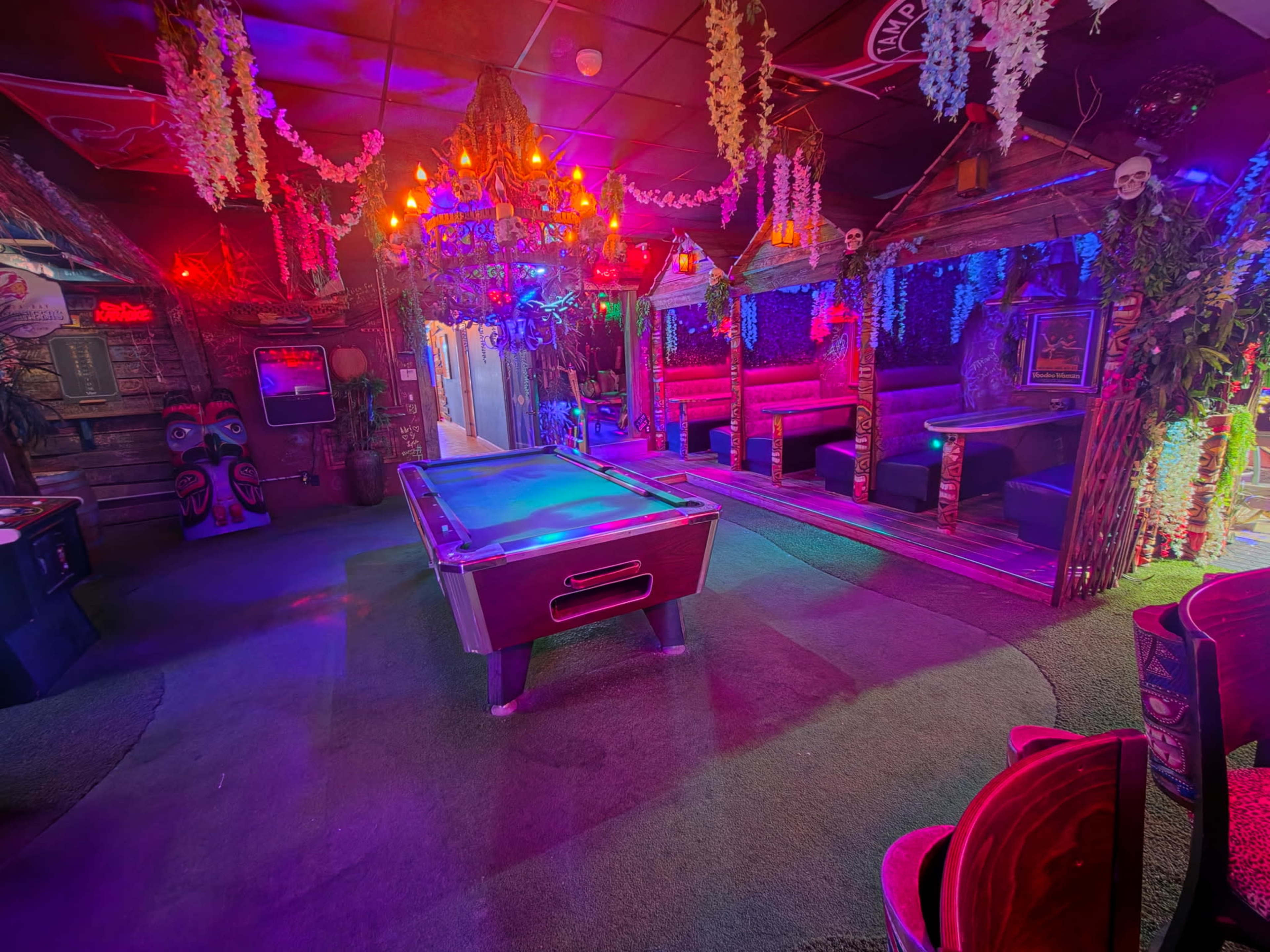 The image shows an illuminated interior of a lively bar or lounge featuring a pool table at the center, surrounded by colorful lighting, seating areas, and decorative elements.