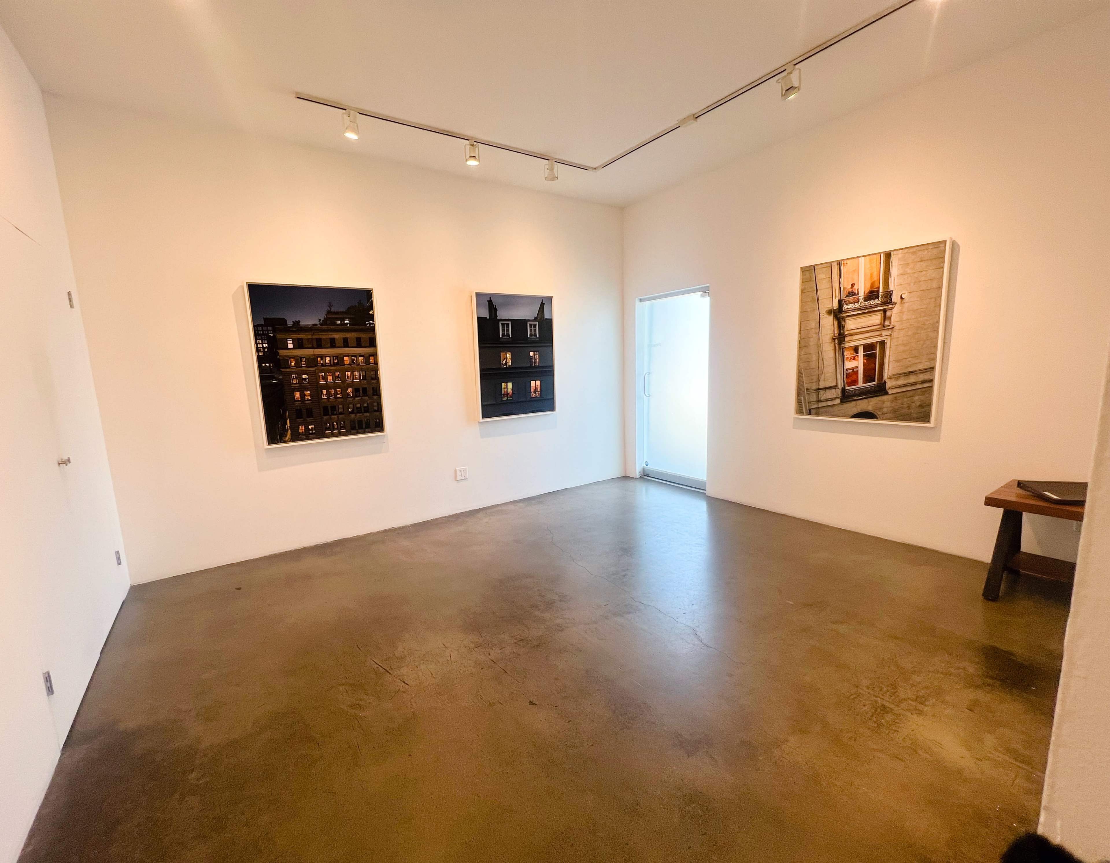 The image shows a modern gallery space with three framed photographs on the white walls and a door leading outside.