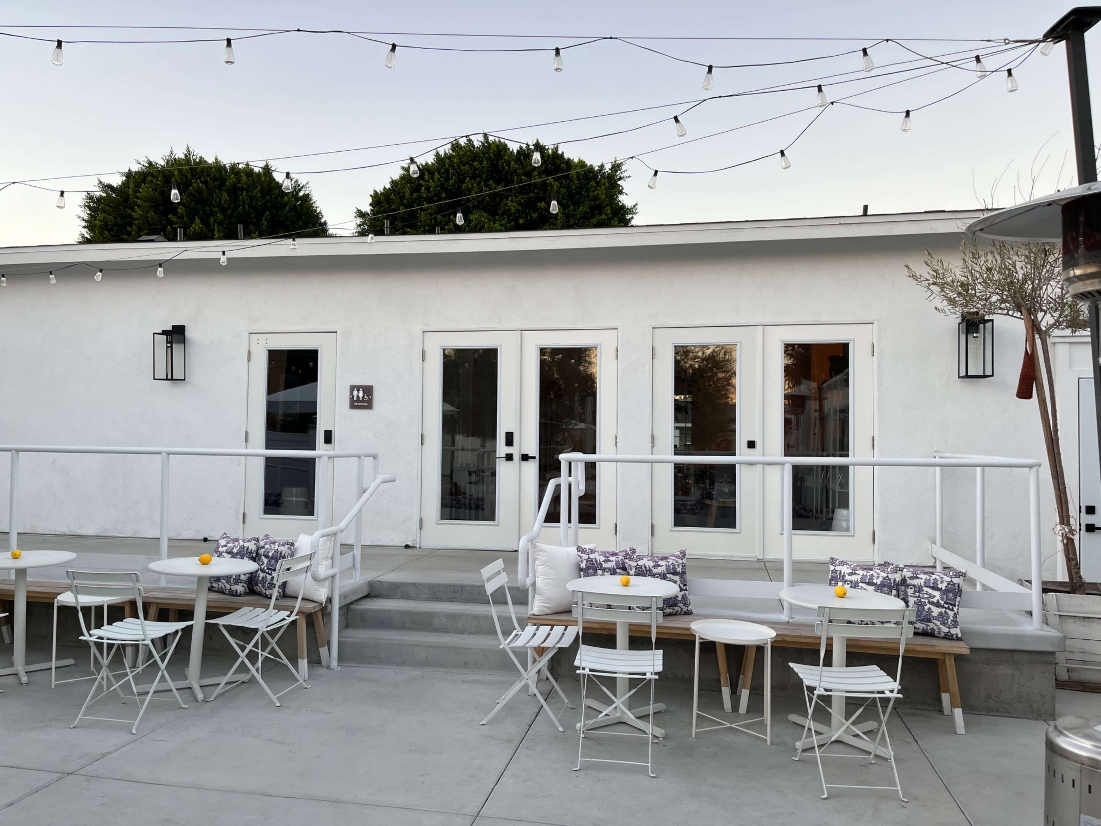 A modern outdoor patio area with white furniture and string lights illuminating a building with glass doors and a minimalist design.