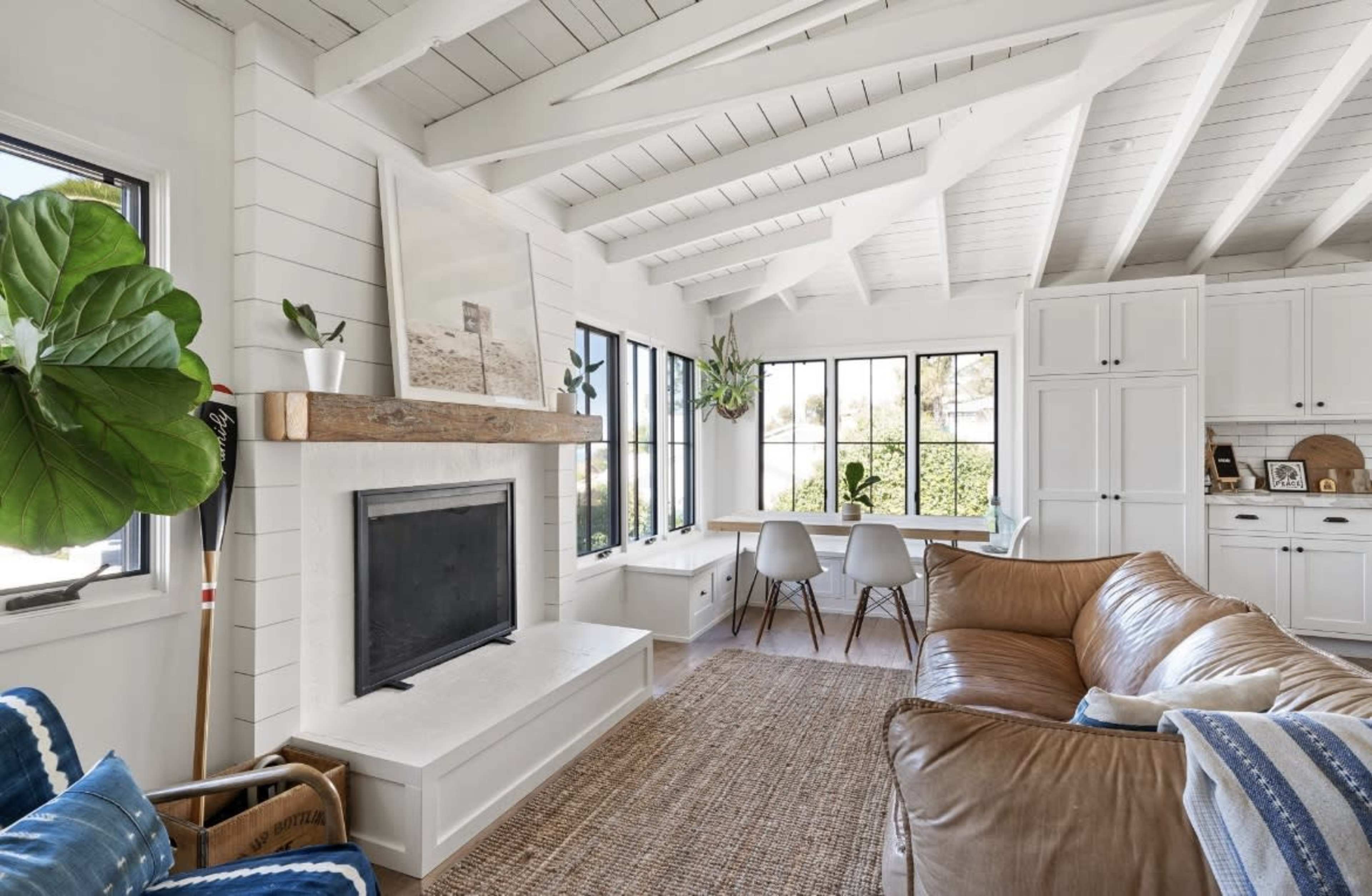 A bright, airy living room with a white ceiling, a fireplace, large windows, a brown leather sofa, and a dining area with a wooden table and chairs.