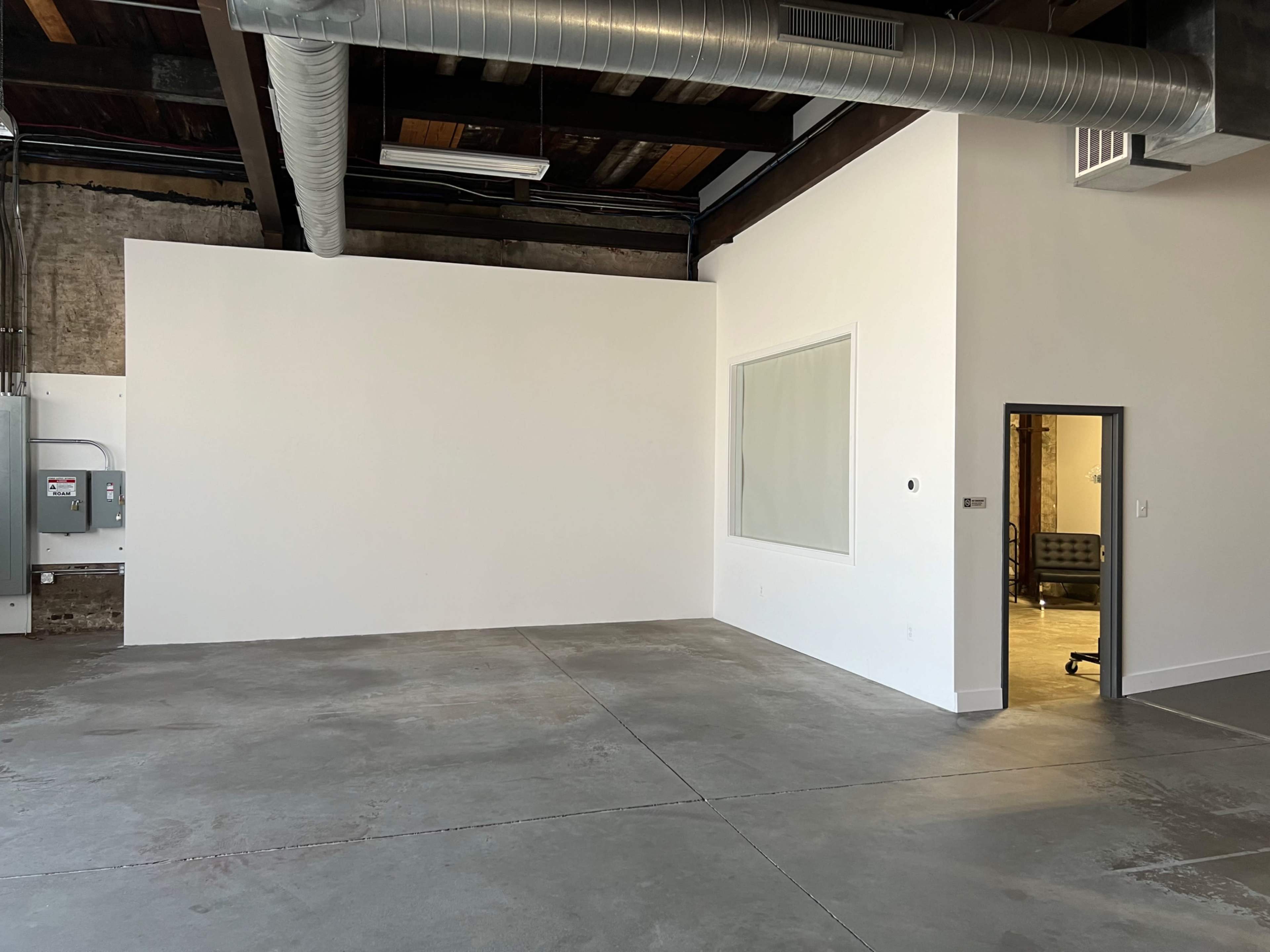 An empty room with a white wall, a large window, and a door leading to another space, set against a concrete floor and exposed ceiling.