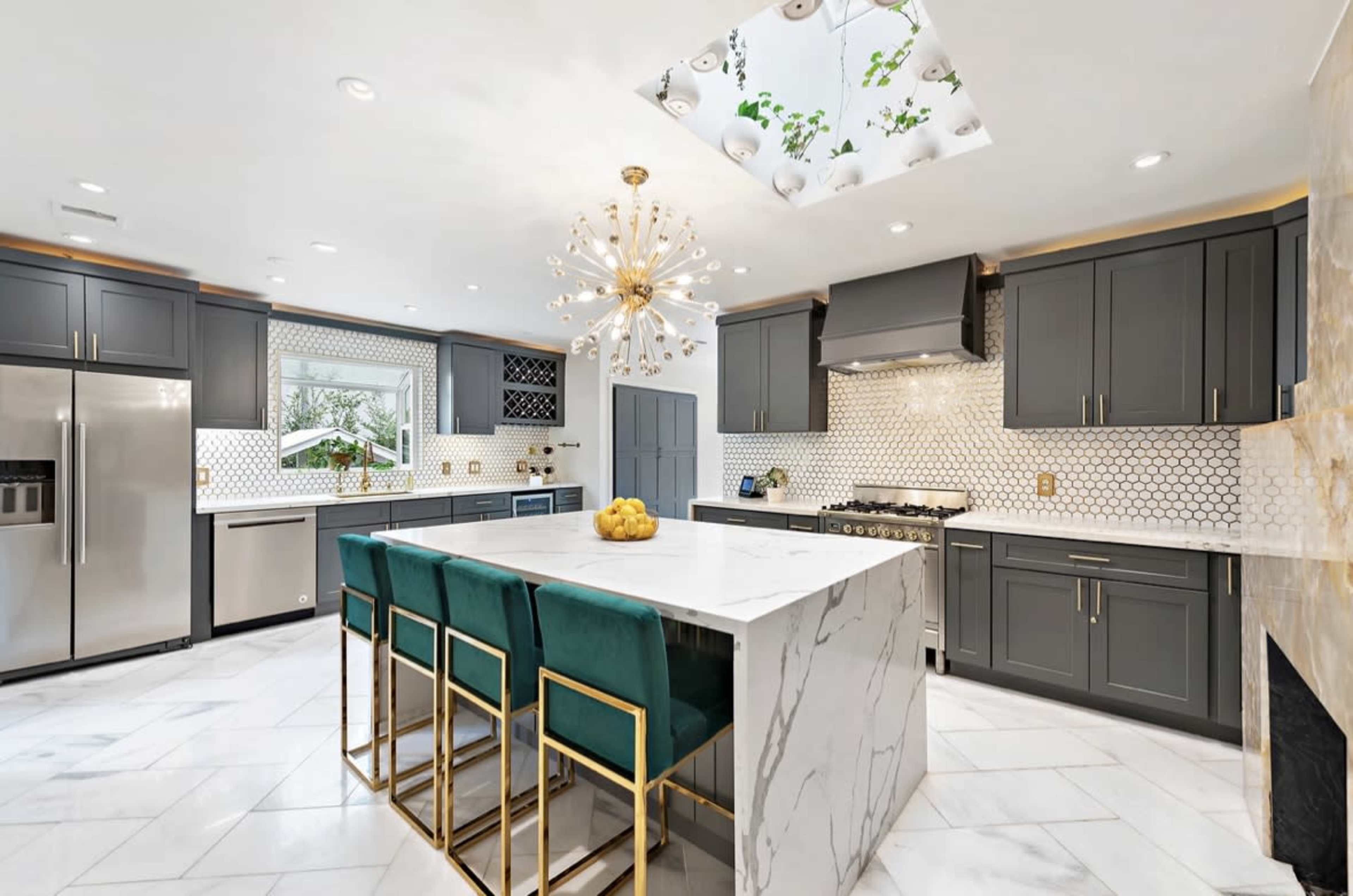 A modern kitchen features gray cabinetry, a large marble island with gold bar stools, and a decorative ceiling that allows light to enter.