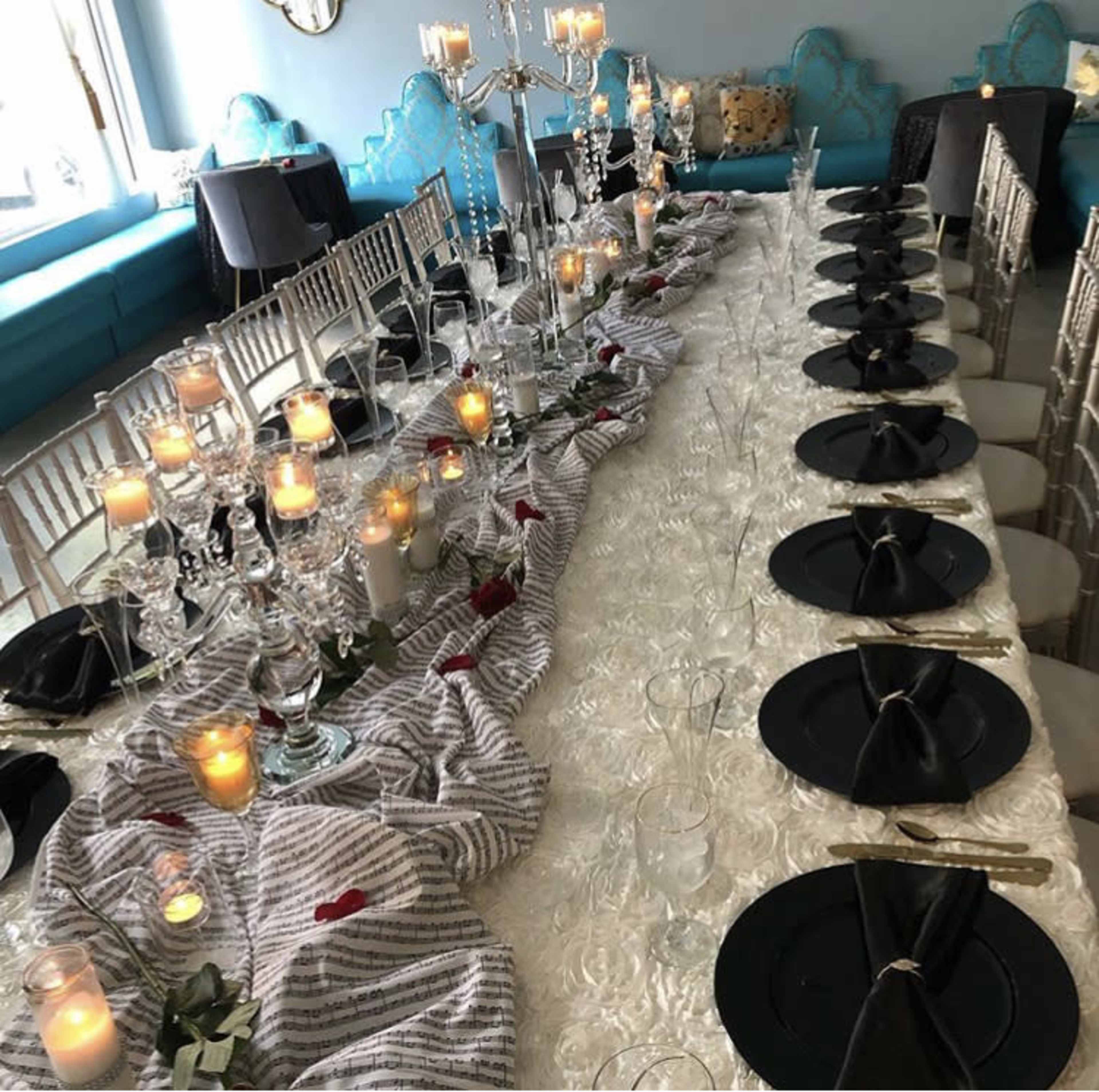 A long dining table is set with black plates, glassware, and candles, adorned with a striped cloth and decorative centerpieces.