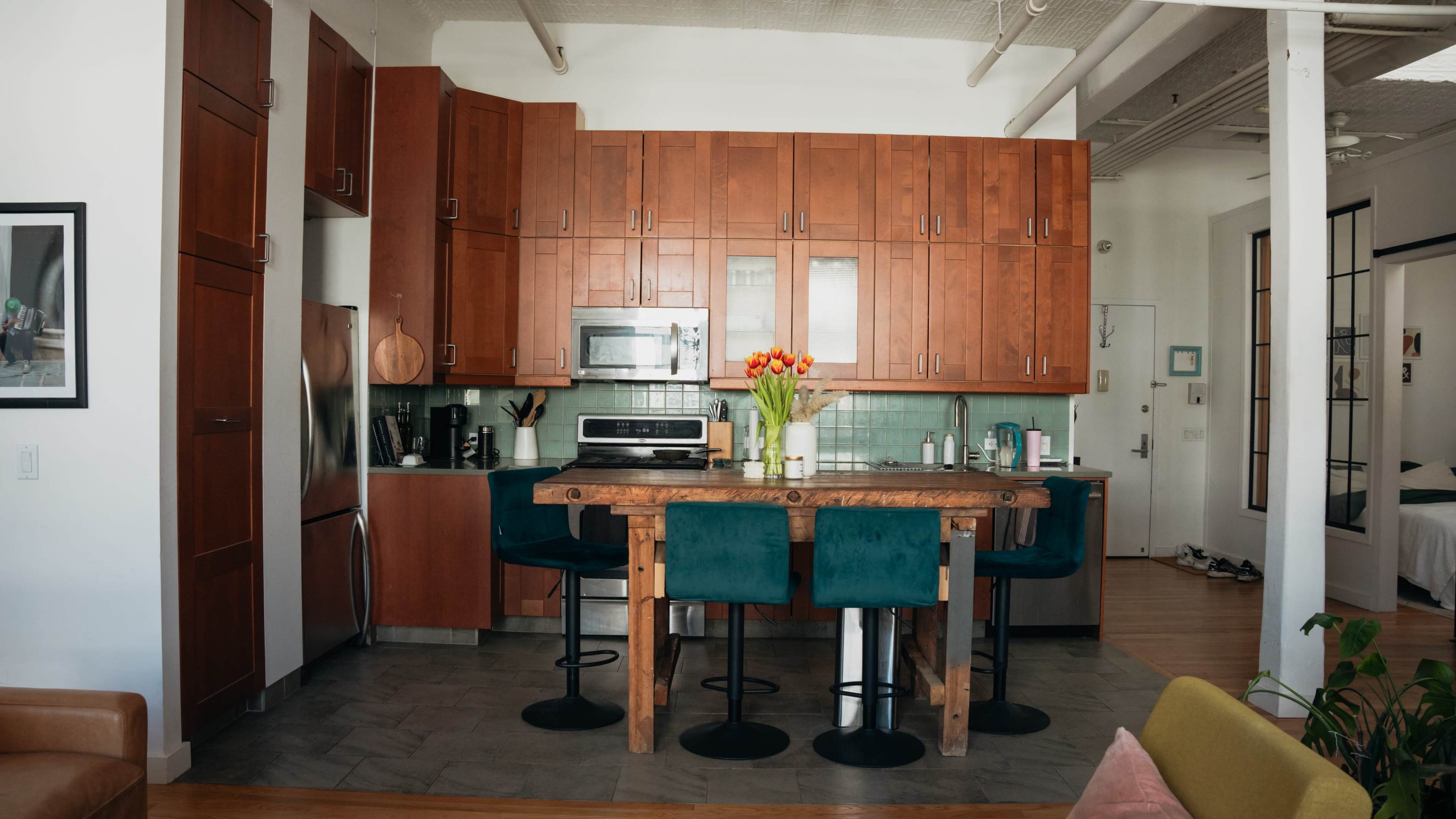 The image shows a modern kitchen with wooden cabinets, a stainless steel refrigerator, and a central wooden table surrounded by teal bar stools.