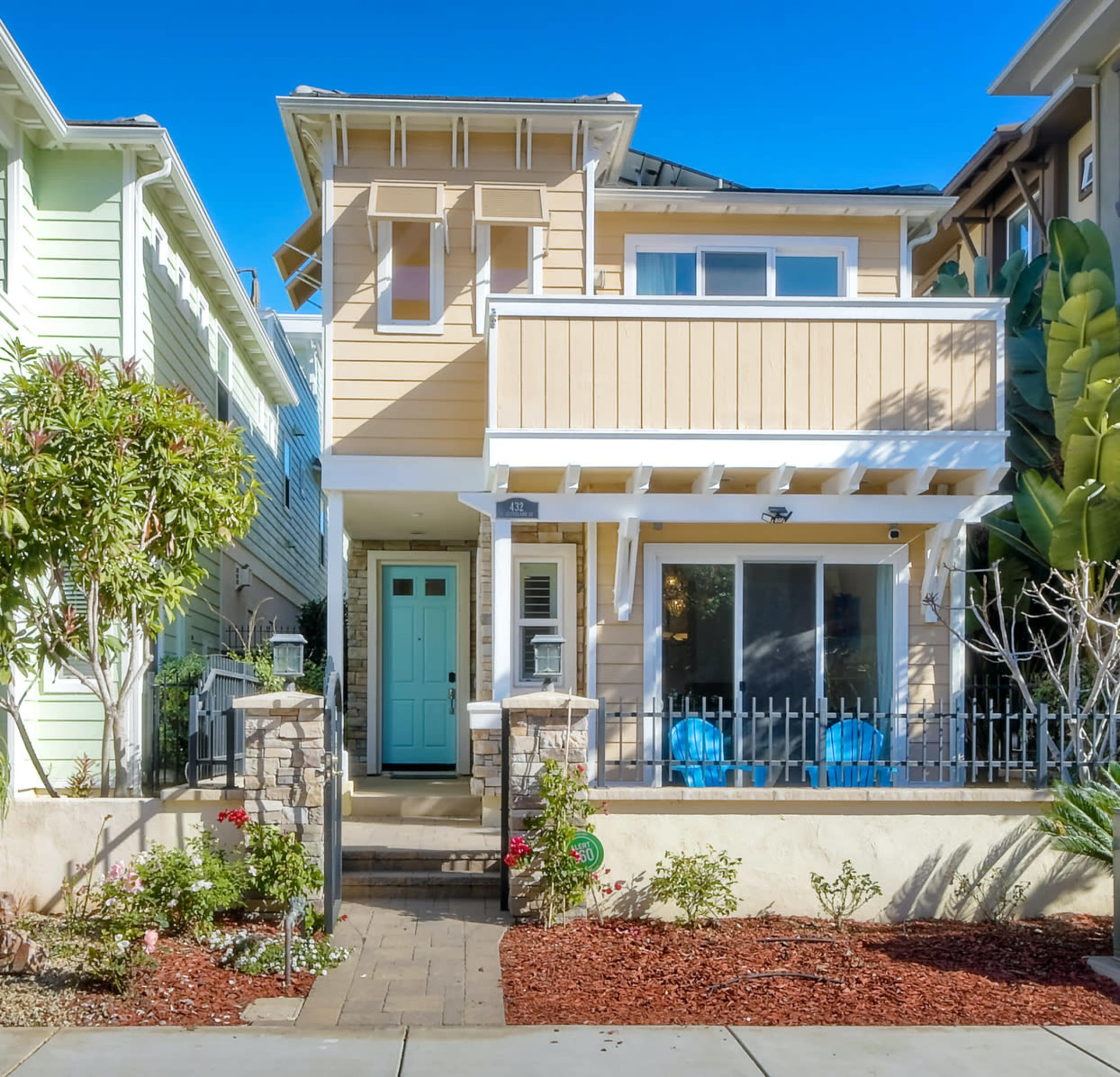 The image shows a two-story house with a pale yellow exterior, a front porch, and a green door, located in a neighborhood with similar homes.