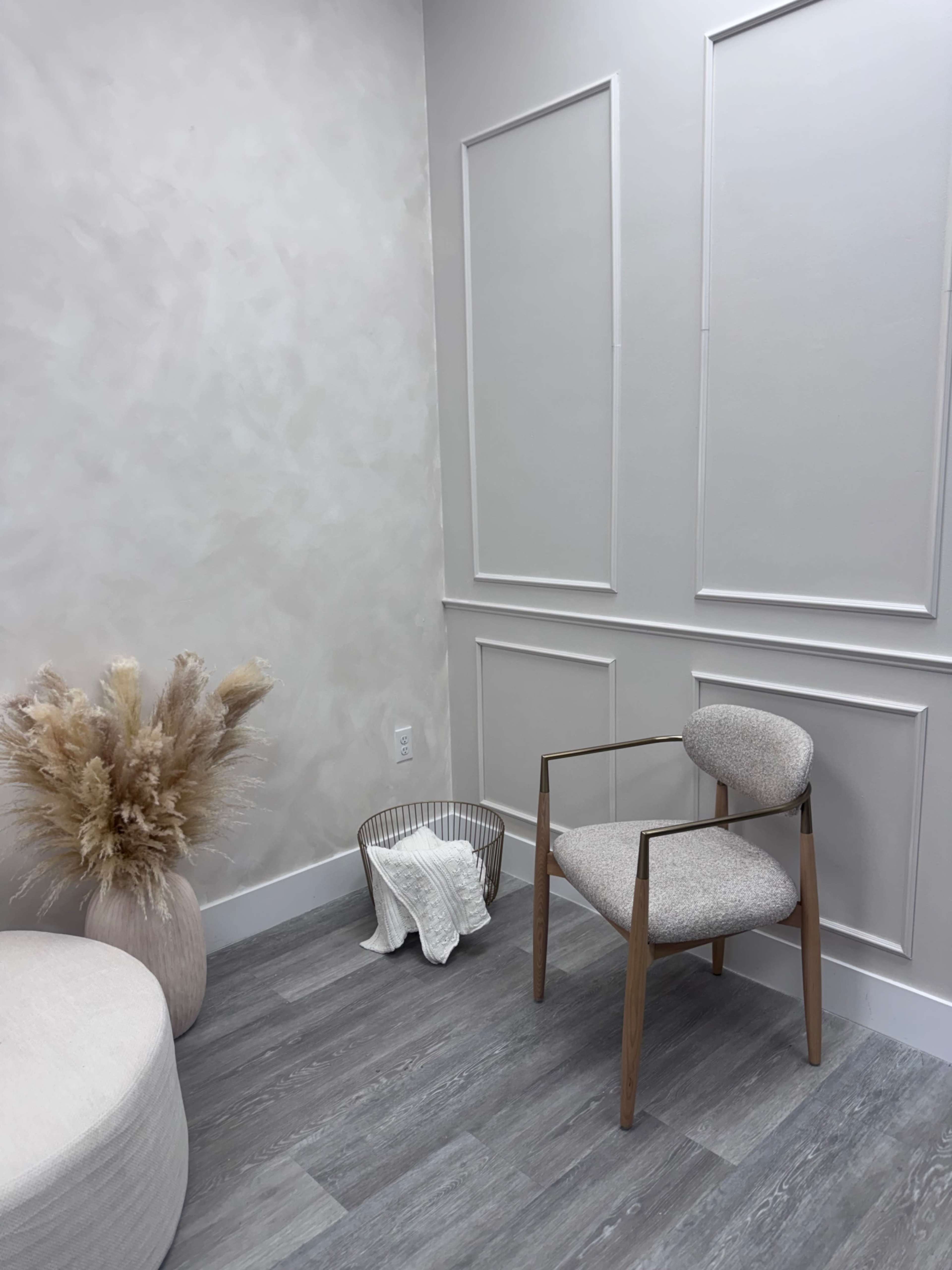 The image shows a minimalist room with a light-colored wall, a wooden chair, a round ottoman, a decorative vase with dried plants, and a metal basket.