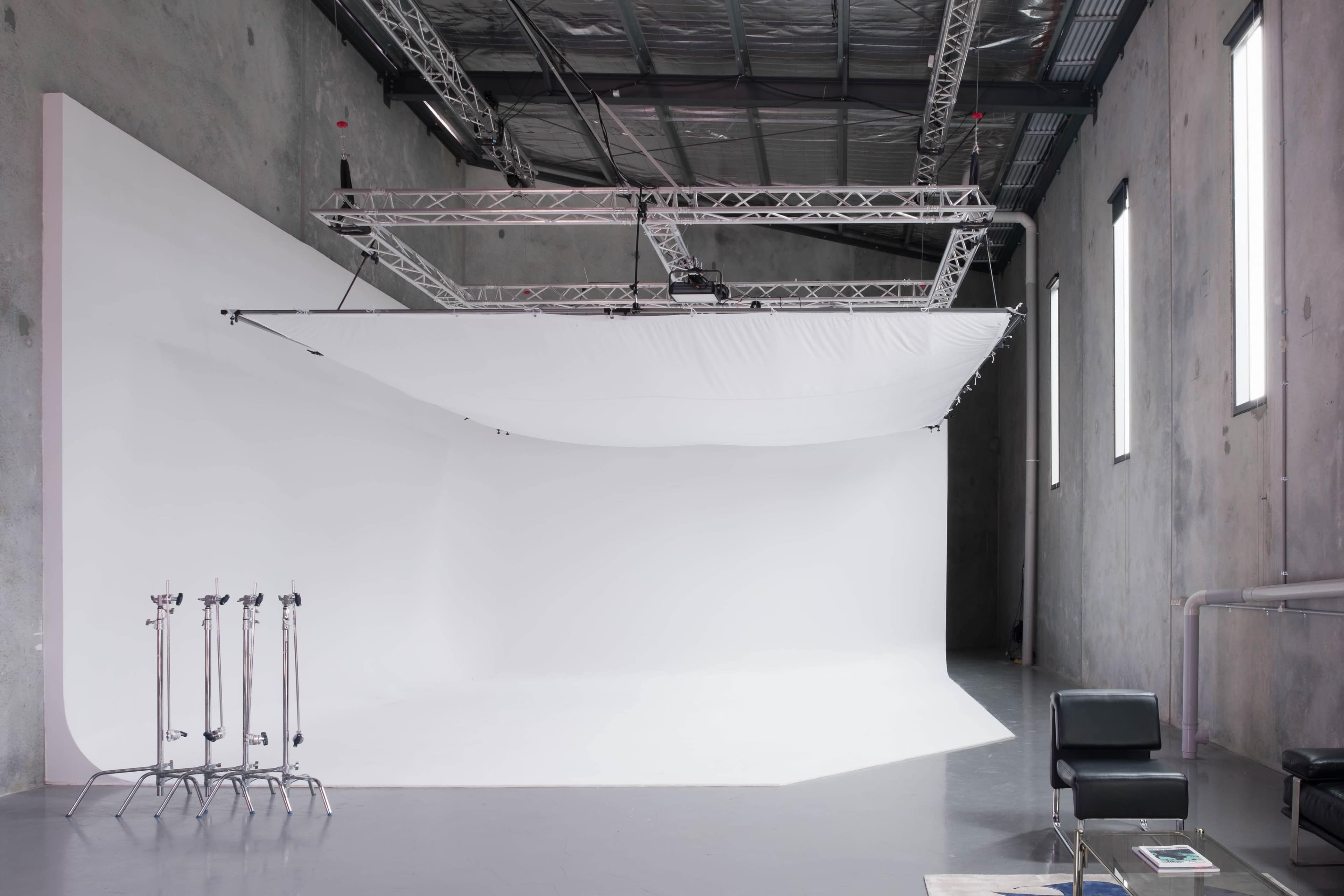 A photography studio with a large white backdrop, metal support structure overhead, and a black chair positioned near the edge.