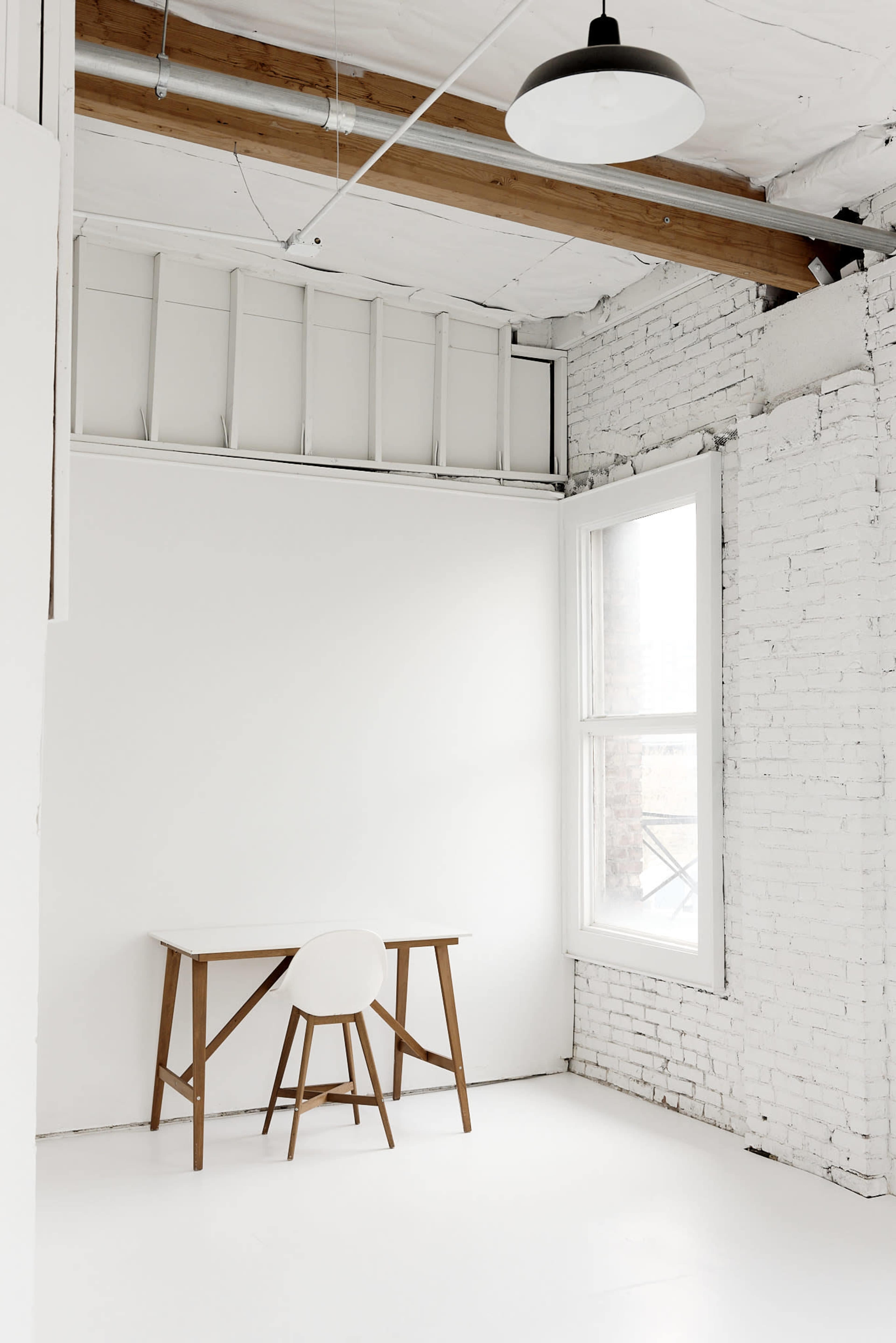 The image shows a minimalist interior space featuring a wooden table and a stool against a white brick wall with large windows.
