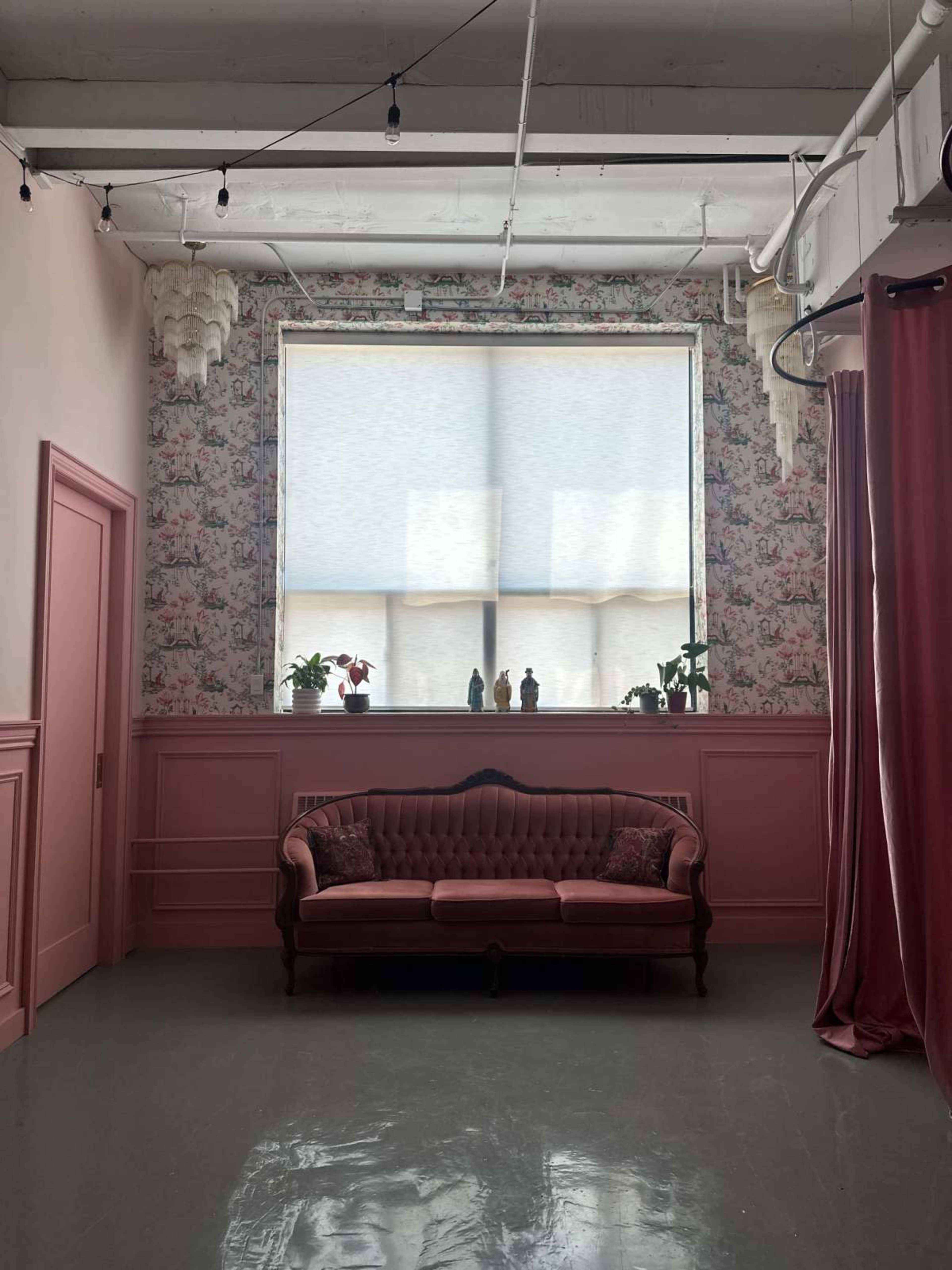 The interior features a pink satin couch against a floral wallpaper backdrop, with a large window covered by light shades and potted plants adorning the sill.