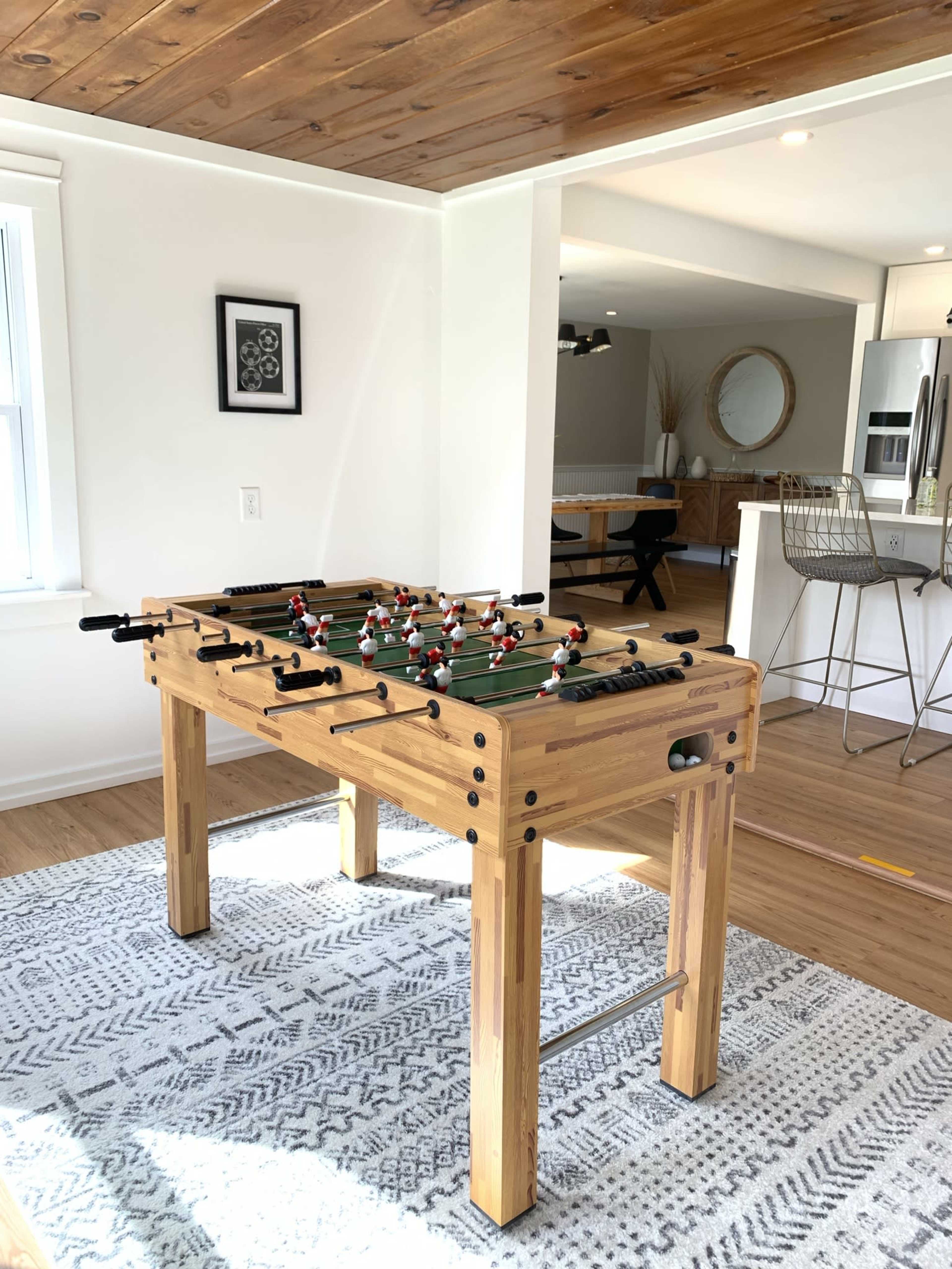 A foosball table sits on a patterned rug in a well-lit room, with a dining area visible in the background.