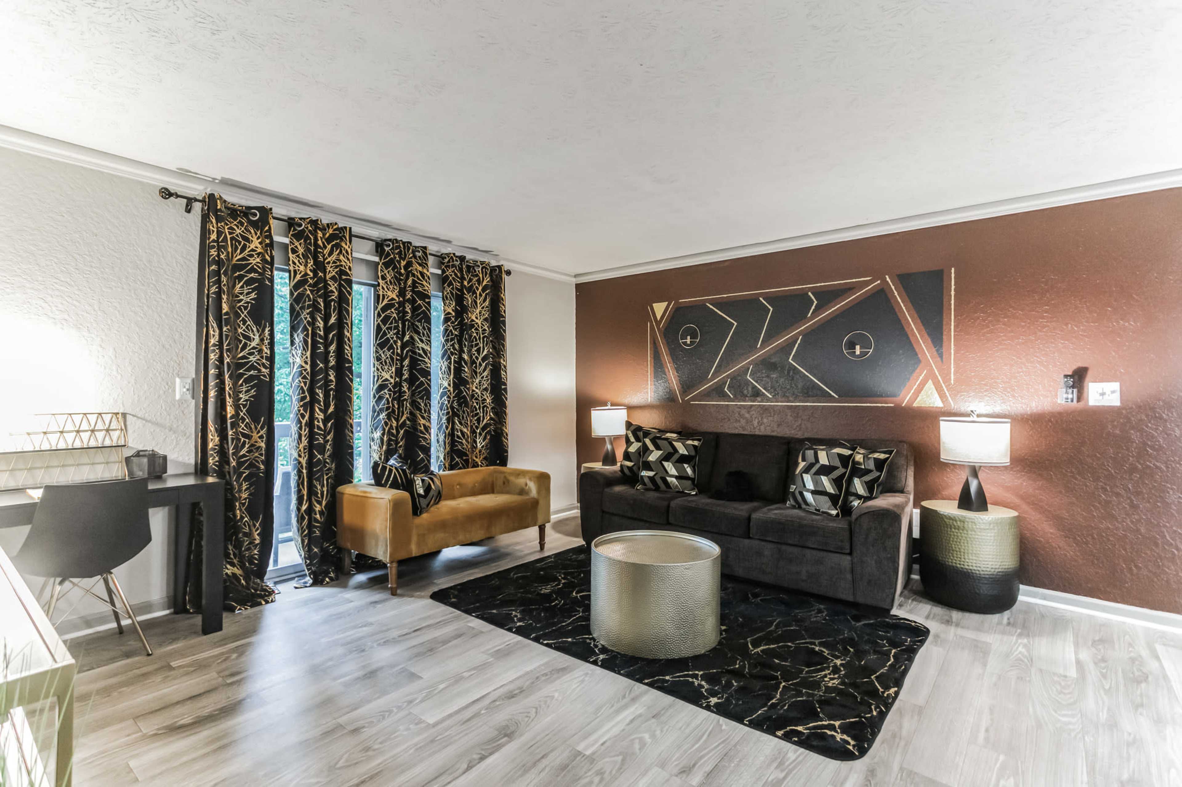 The scene shows a modern living room featuring a dark couch with decorative pillows, an accent chair, a round coffee table, patterned curtains, and a painting on the wall.