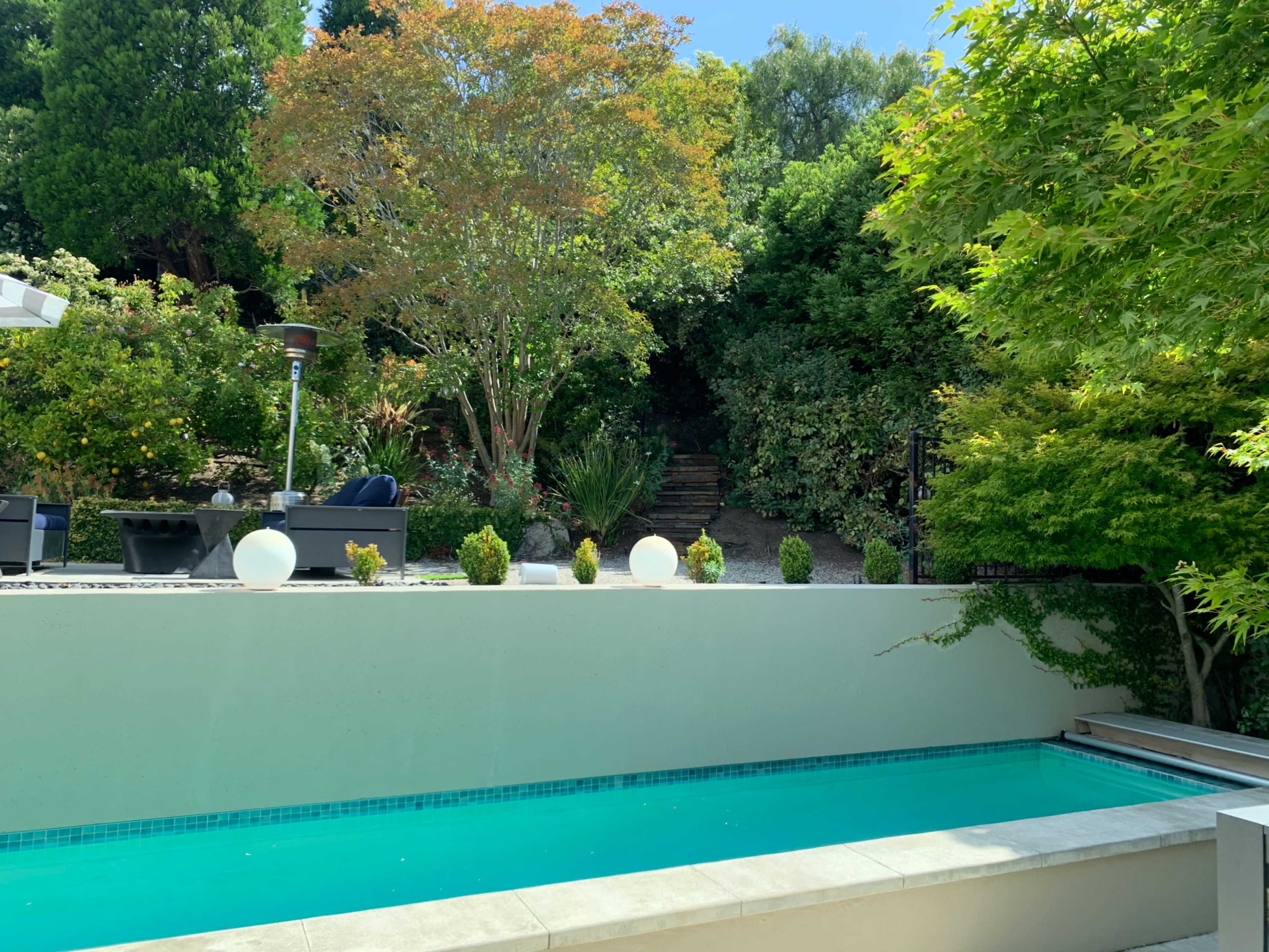 A serene backyard with a turquoise pool, lush greenery, and stone steps leading up to a landscaped area.