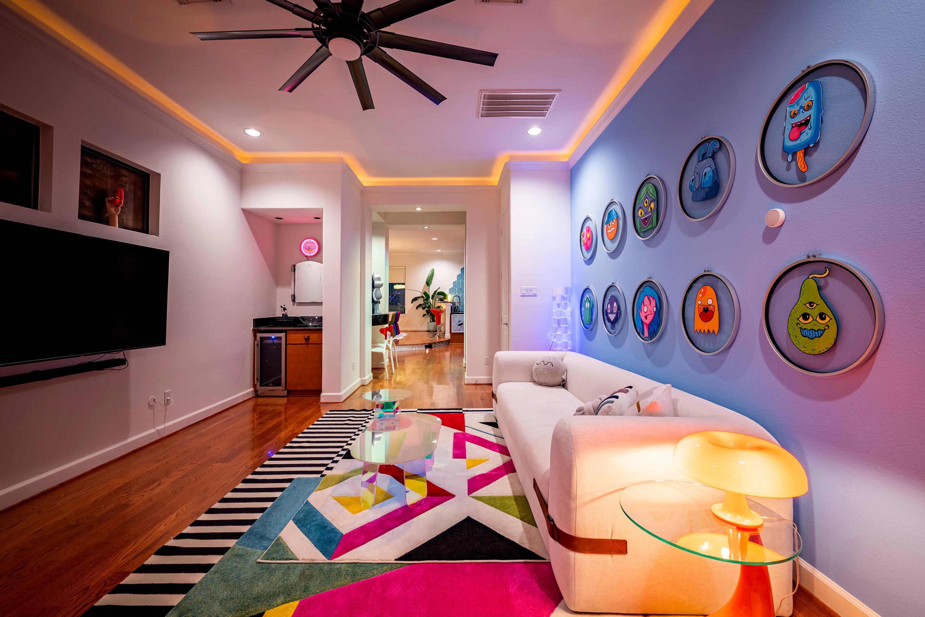 The image shows a modern living room with a white sofa, colorful patterned rug, wall-mounted artwork, and a kitchen area visible in the background.