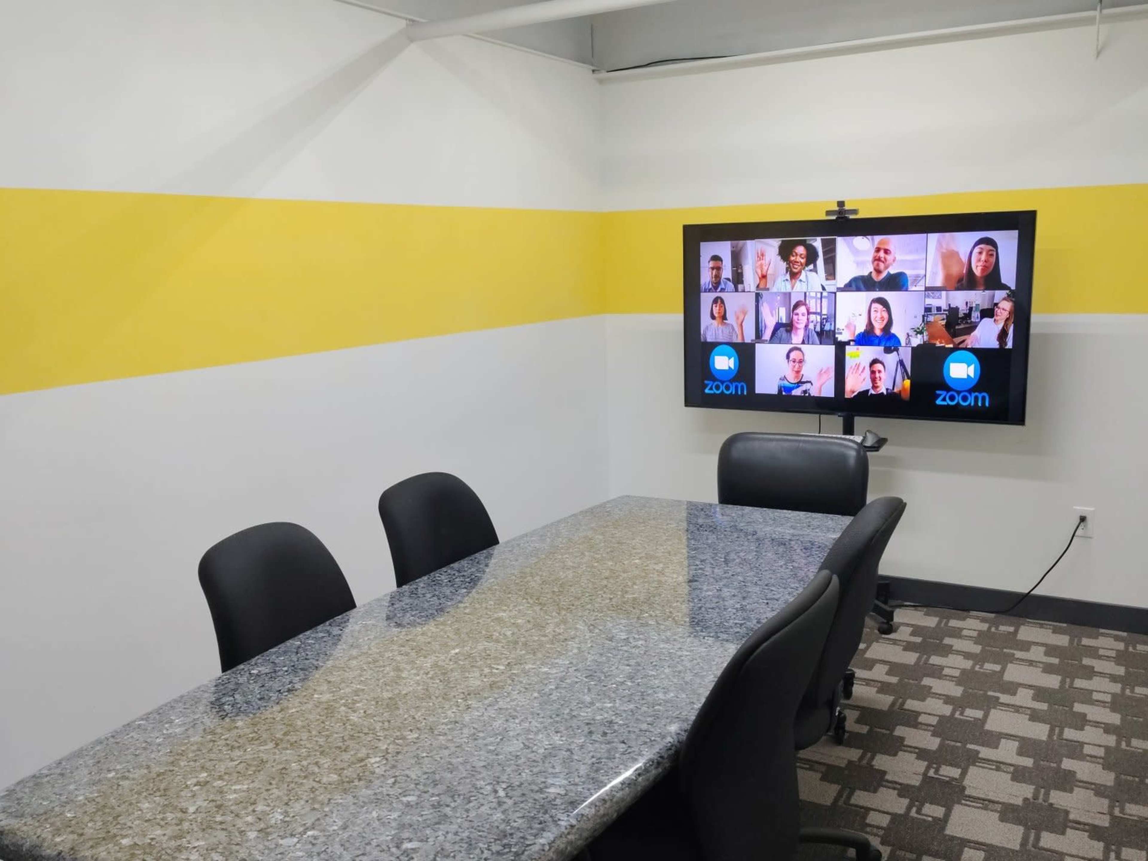 A conference room features a large gray table surrounded by black chairs and a screen displaying a Zoom video call with multiple participants.