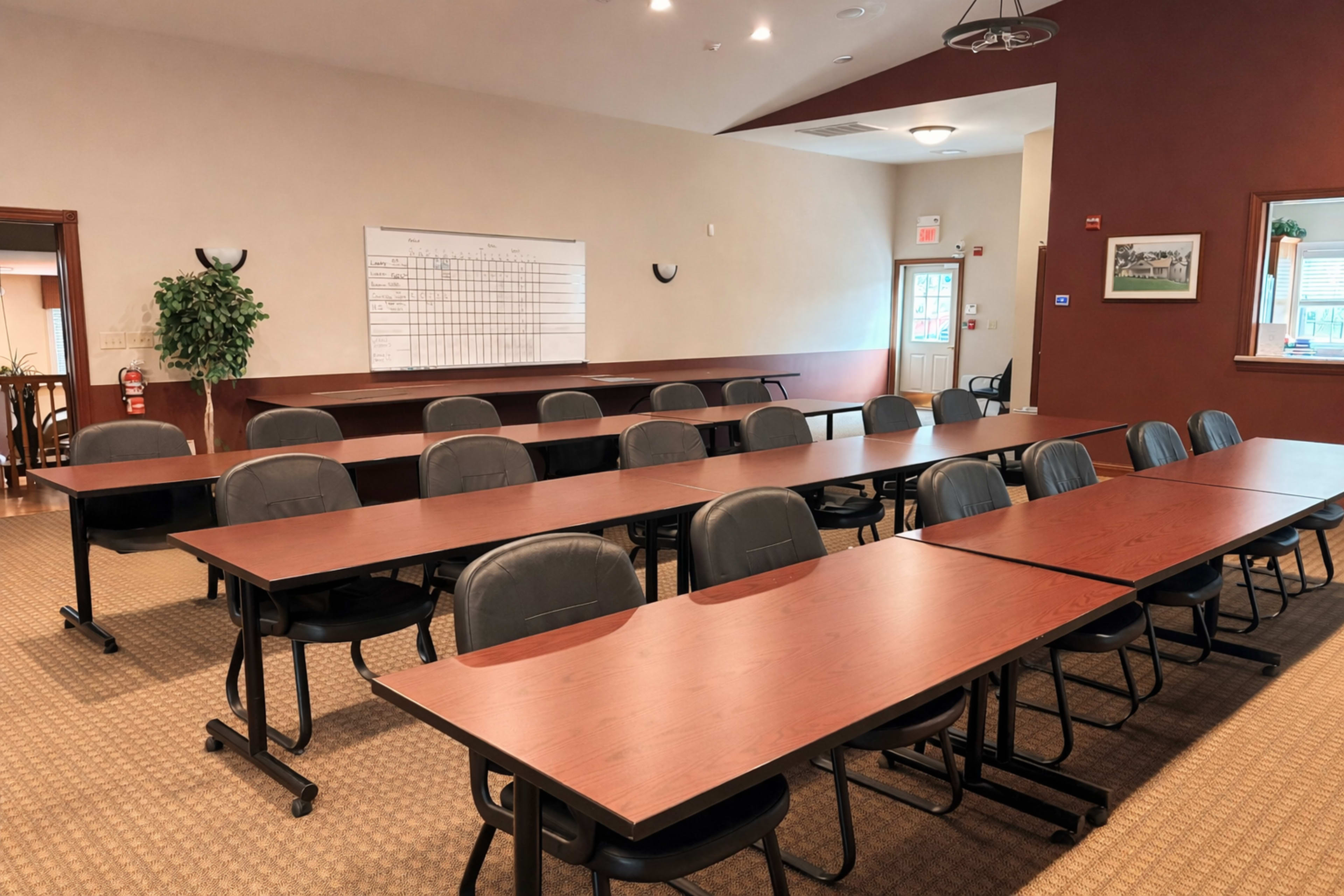 The image shows a meeting room with rows of tables and chairs arranged neatly, facing a wall with a whiteboard.