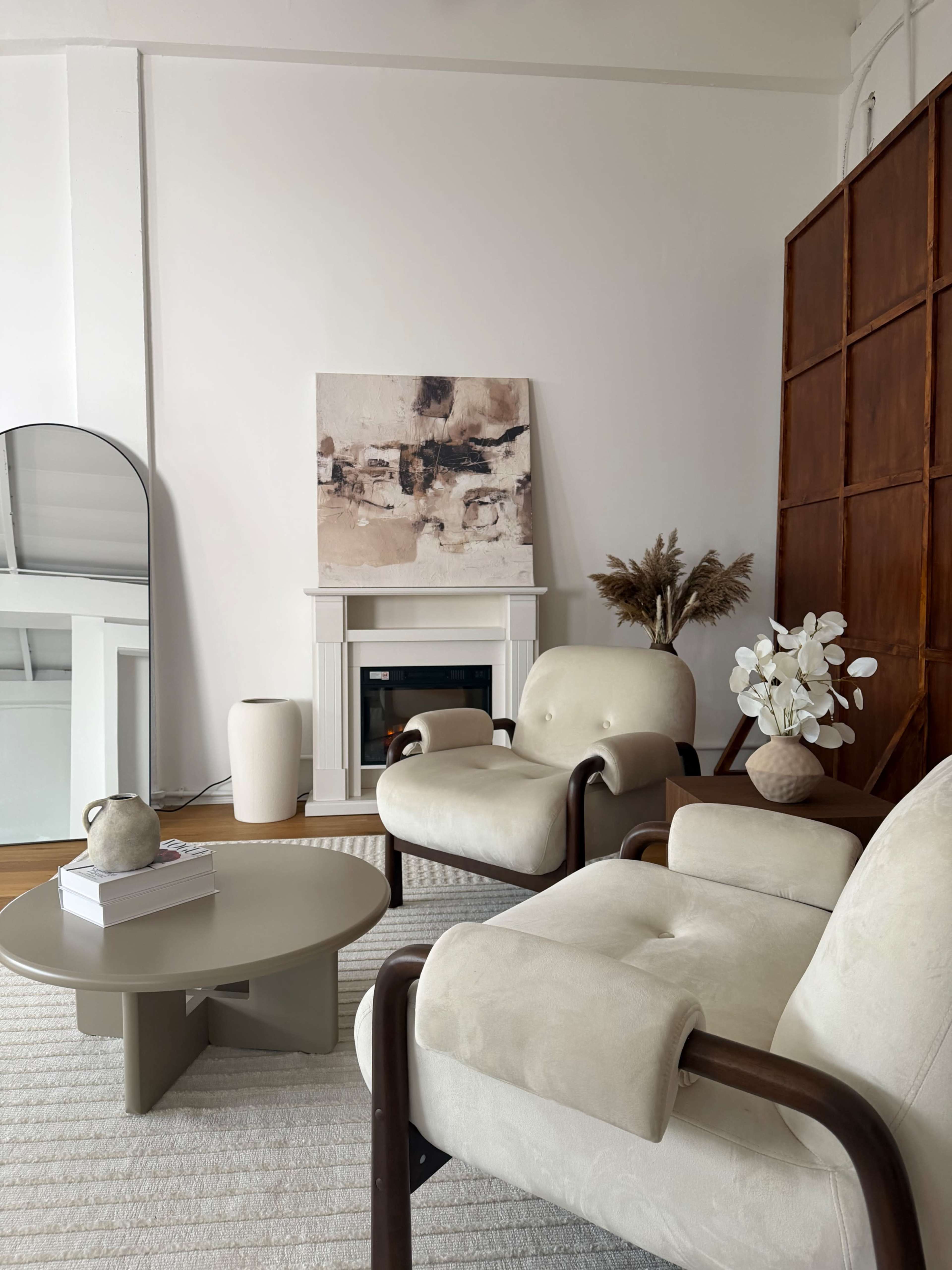 The image depicts a modern living room featuring two upholstered armchairs, a round coffee table, and a minimalist artwork above a fireplace, complemented by decorative elements like vases and plants.