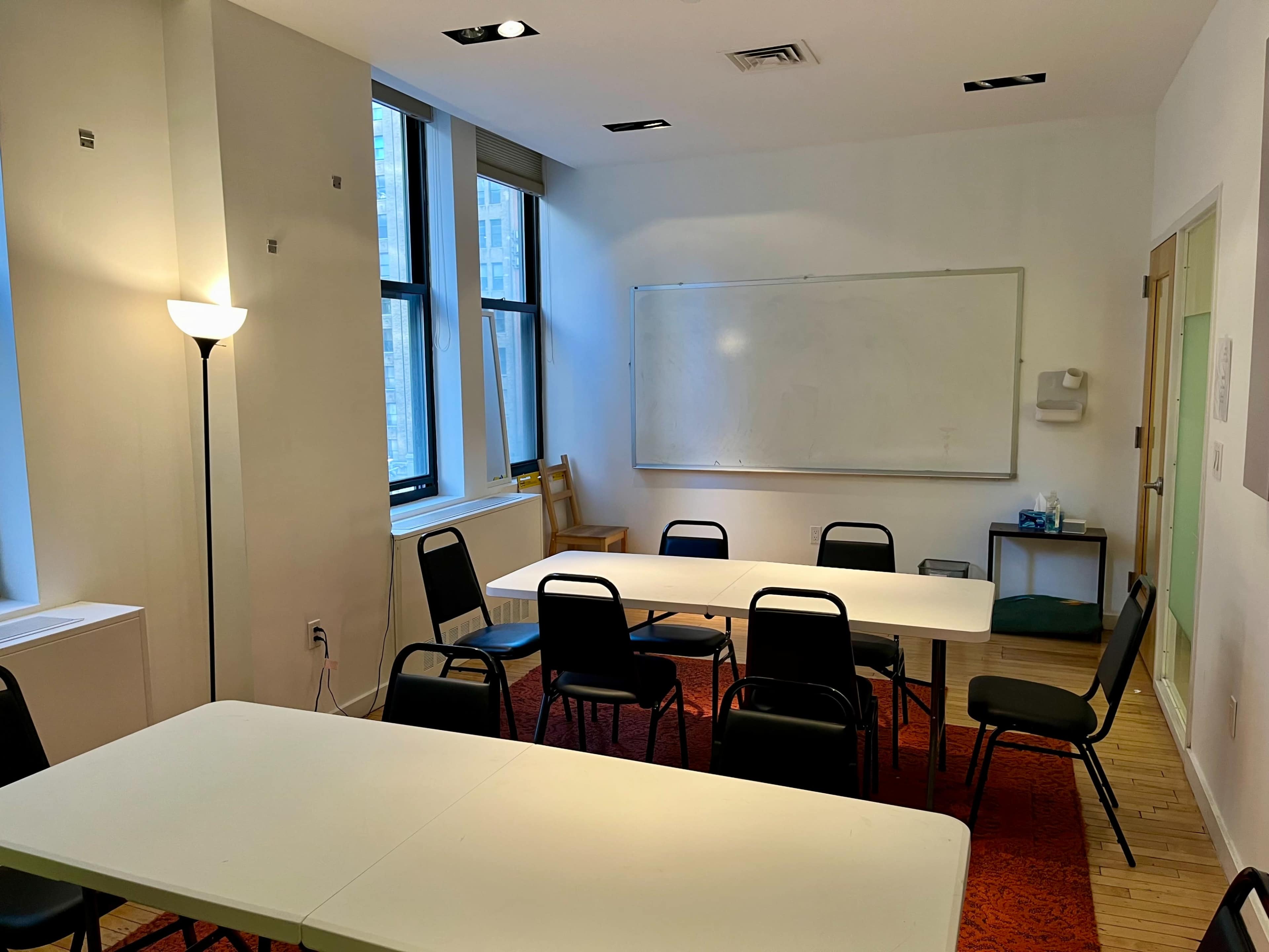 The image shows a sparsely furnished meeting room with a whiteboard, several chairs, and tables, illuminated by natural light from large windows.