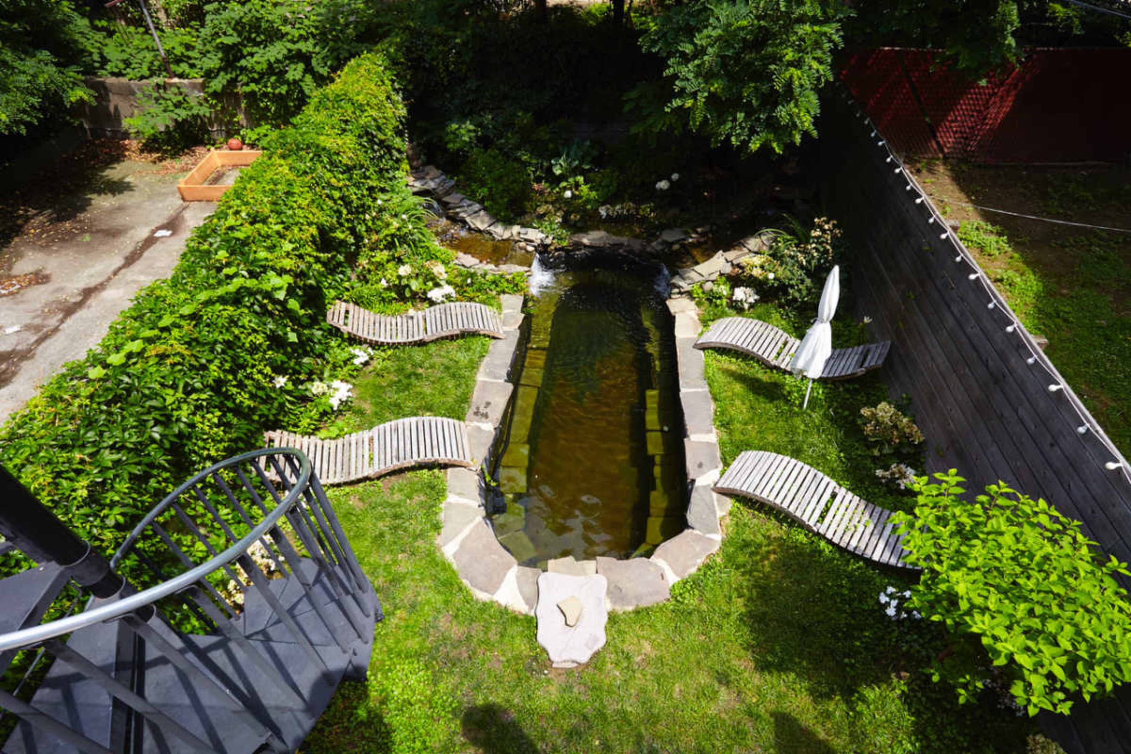 A narrow, stone-lined pond is surrounded by grassy areas and wooden lounge chairs, with lush greenery framing the scene.