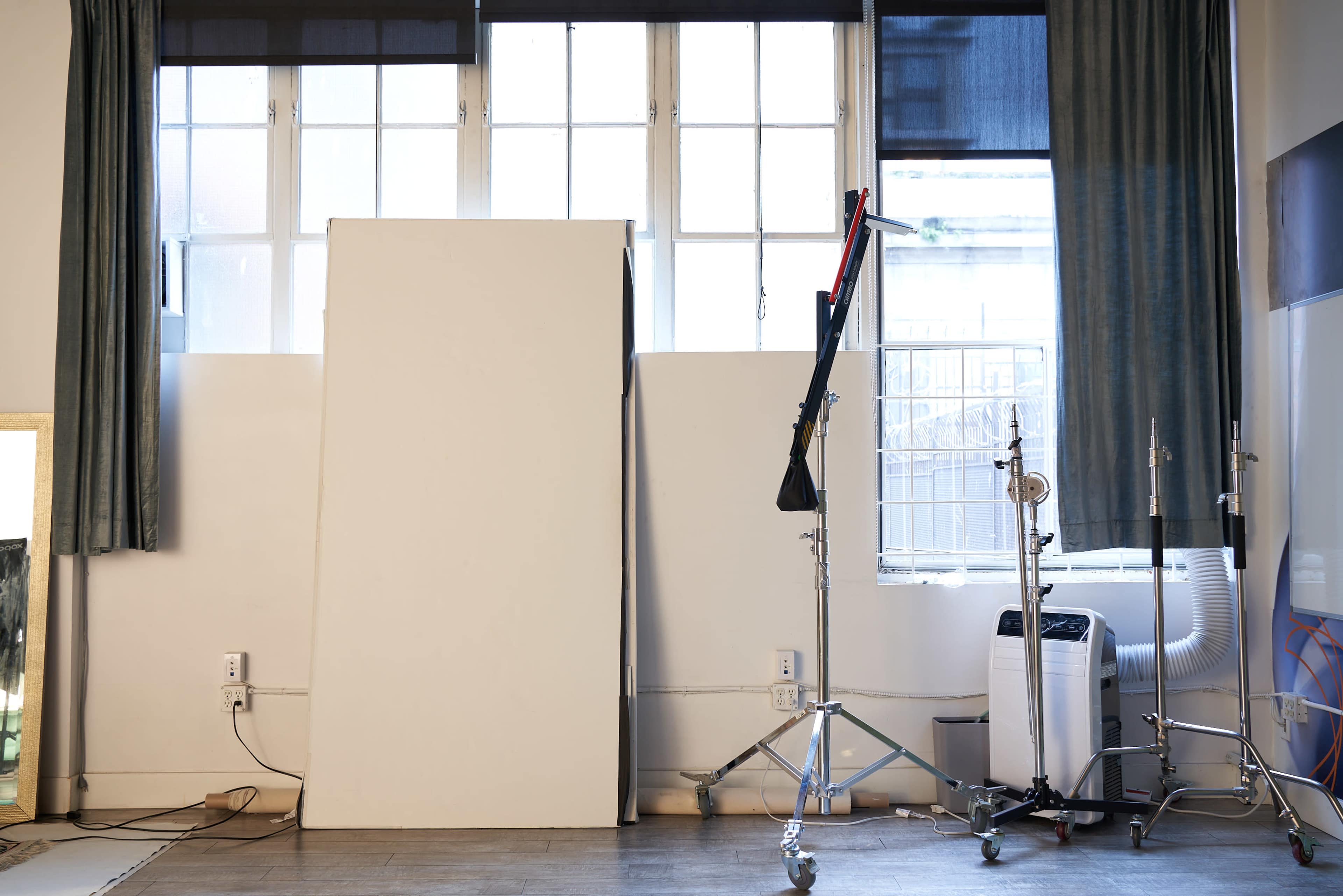The image shows a studio room with large windows, a blank white backdrop leaning against a wall, and a few equipment stands and an air conditioning unit on the floor.