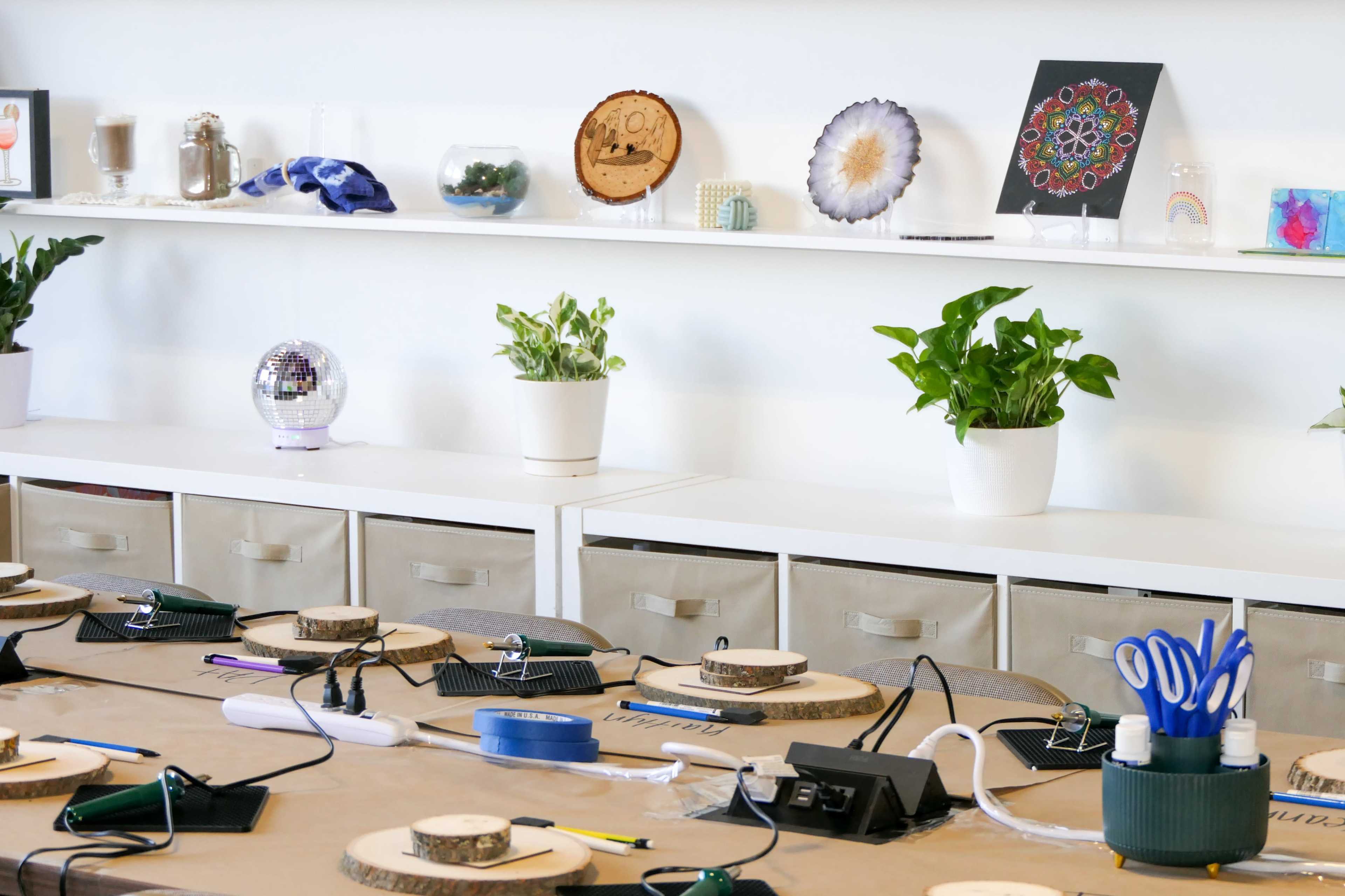 A workspace features a long table with circular wooden bases, surrounded by various tools, while a shelf displays decorative items and potted plants.