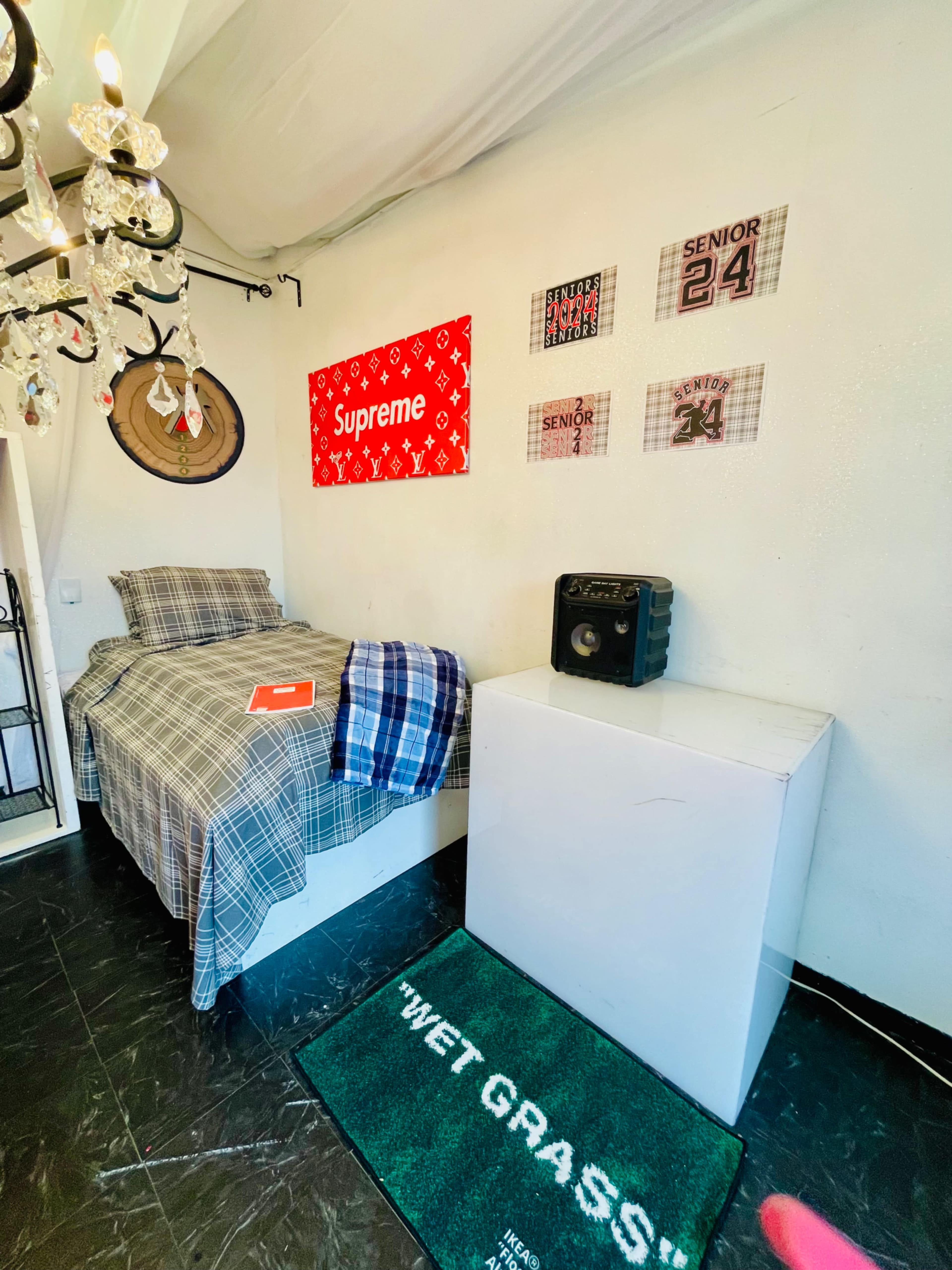 The image shows a small bedroom featuring a bed with a plaid blanket, a white nightstand, a vintage radio, and wall decorations including a "Supreme" poster and framed plaques.