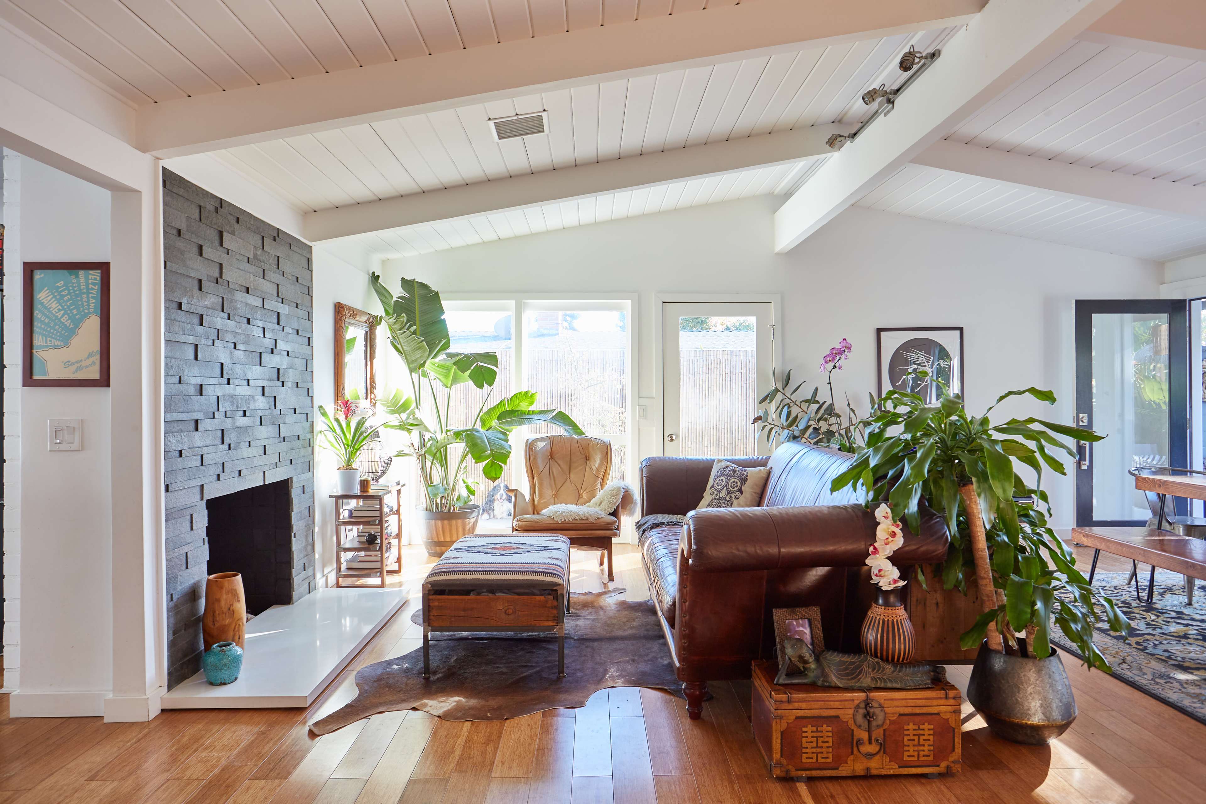 A bright living room features a leather sofa, wooden coffee table, and several potted plants beside a stone fireplace.