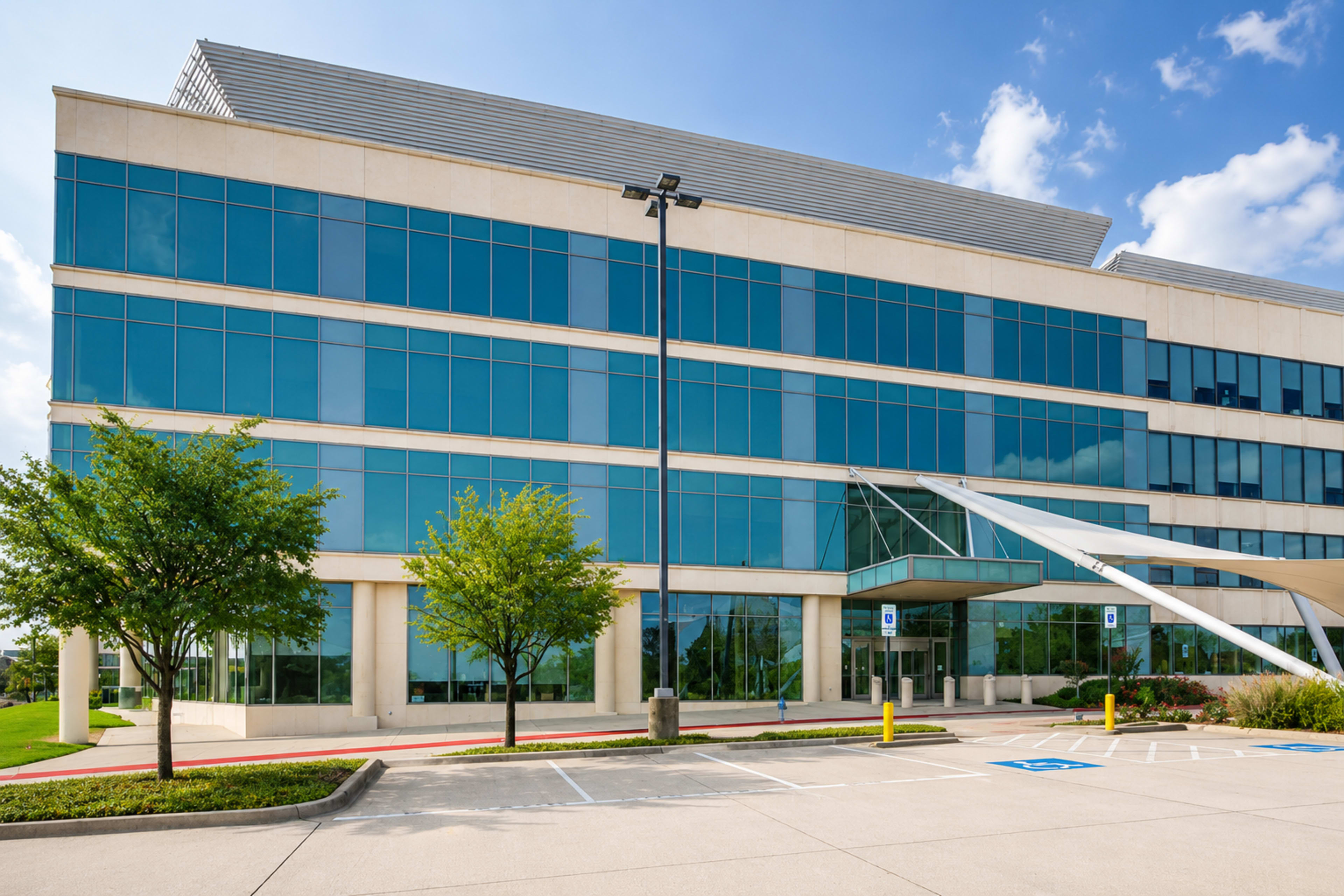 The image shows a modern office building with large glass windows and a landscaped parking area.