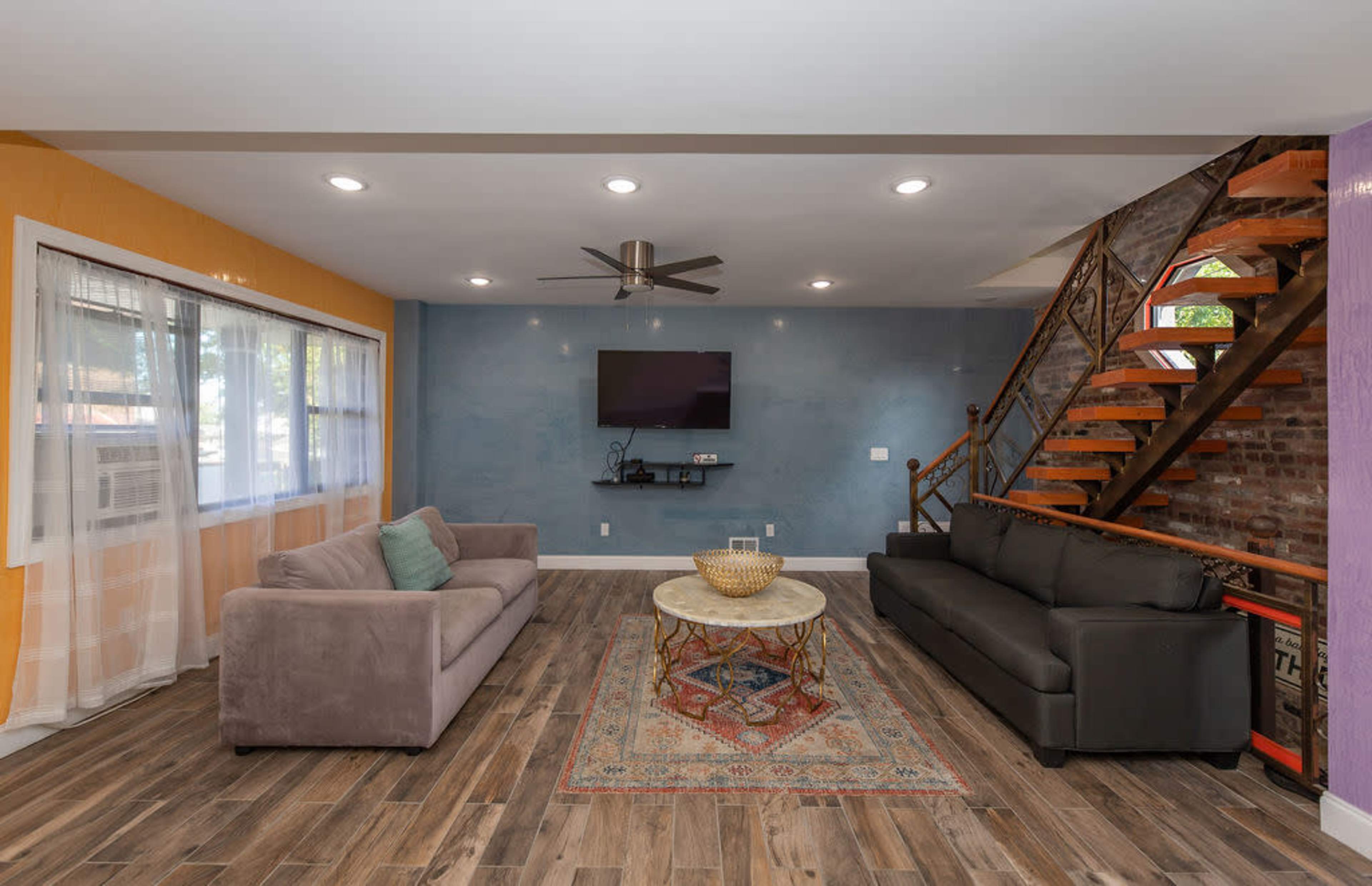 A living room with two sofas, a decorative rug, a coffee table, a wall-mounted TV, and a staircase made of wood and metal.