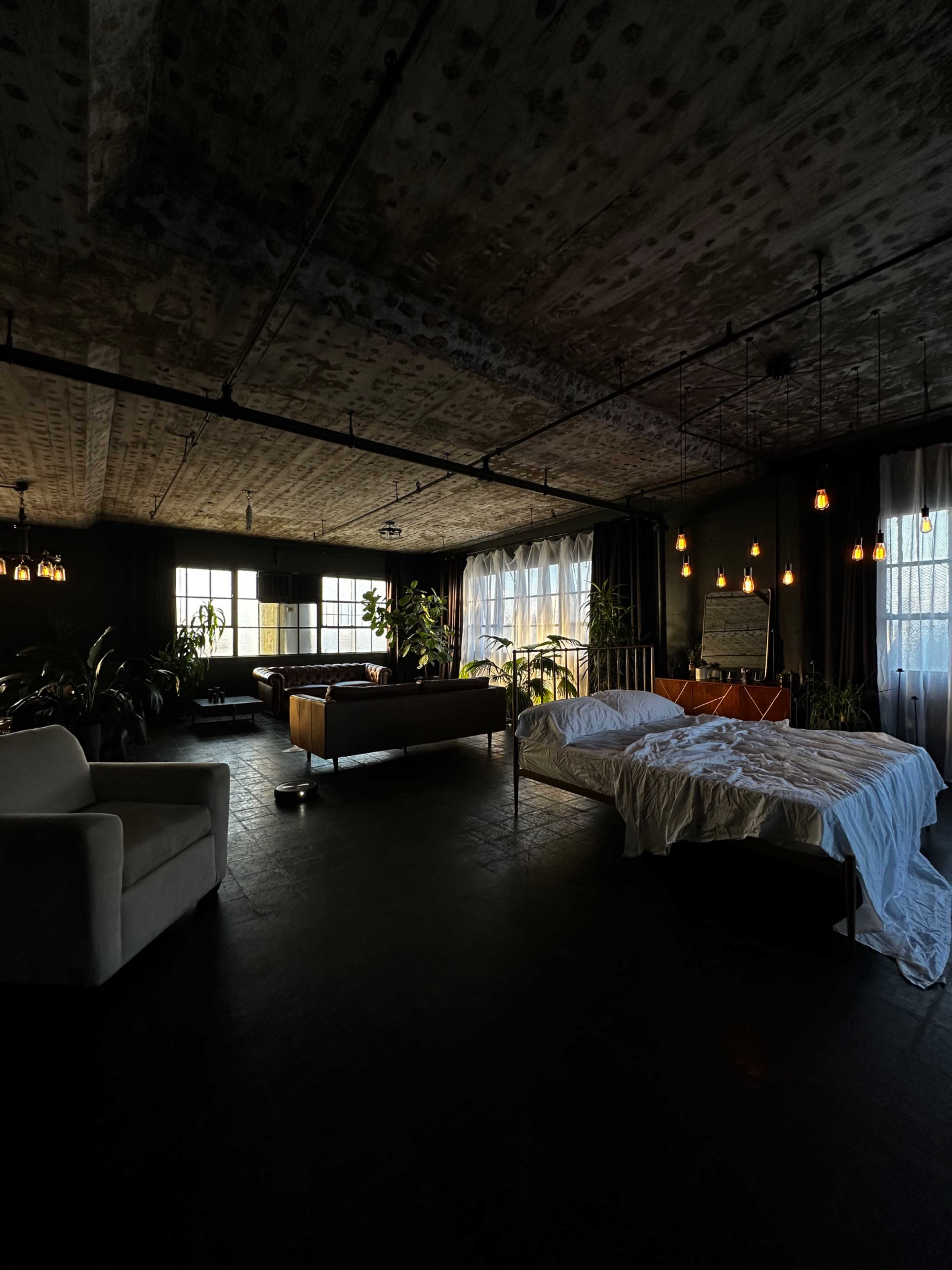 The image shows a spacious interior with a bed draped in white linens, a seating area with a brown couch, large windows, and potted plants throughout.