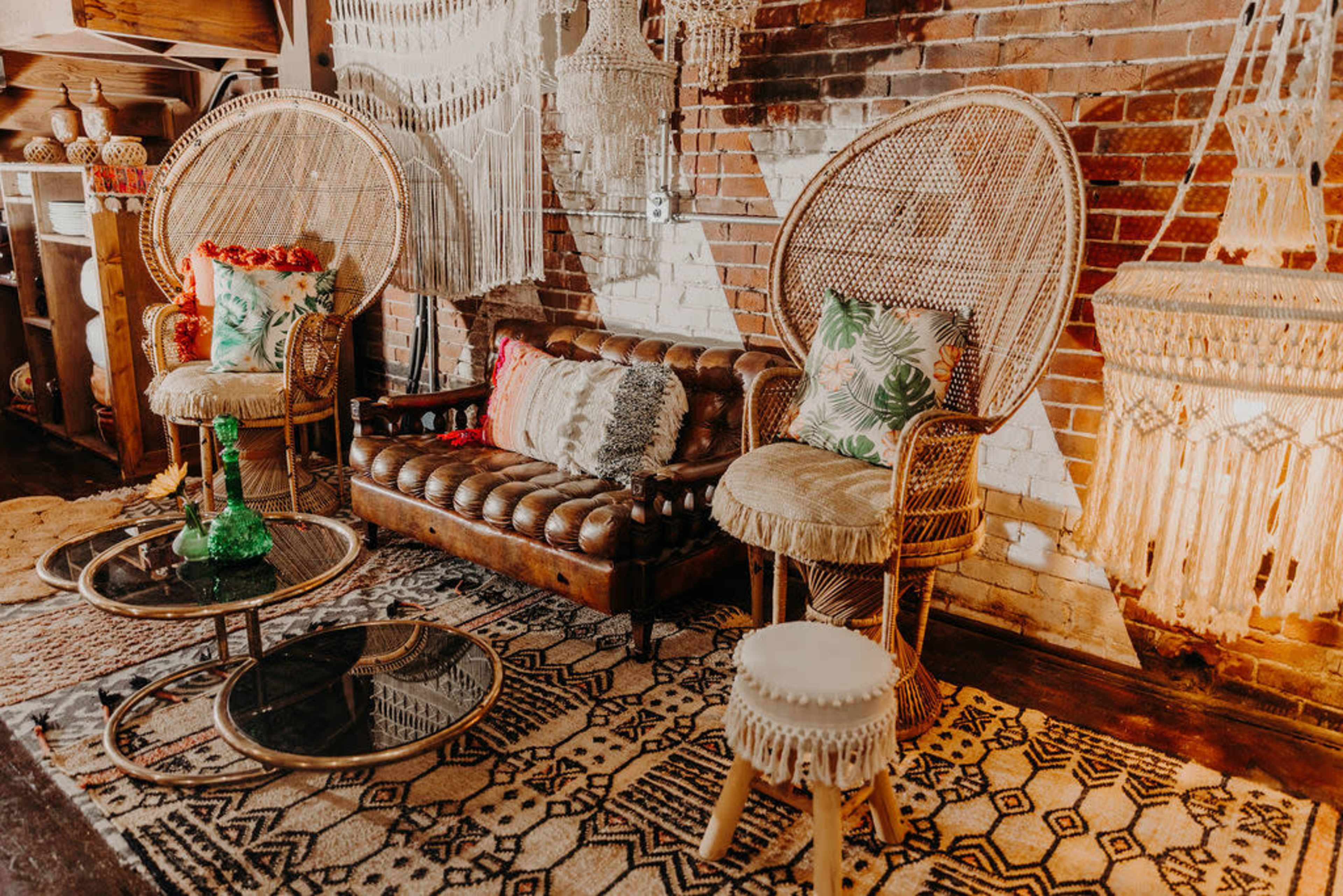A cozy living area featuring a vintage leather sofa, two large wicker chairs, and decorative tables on a patterned rug against a brick wall.