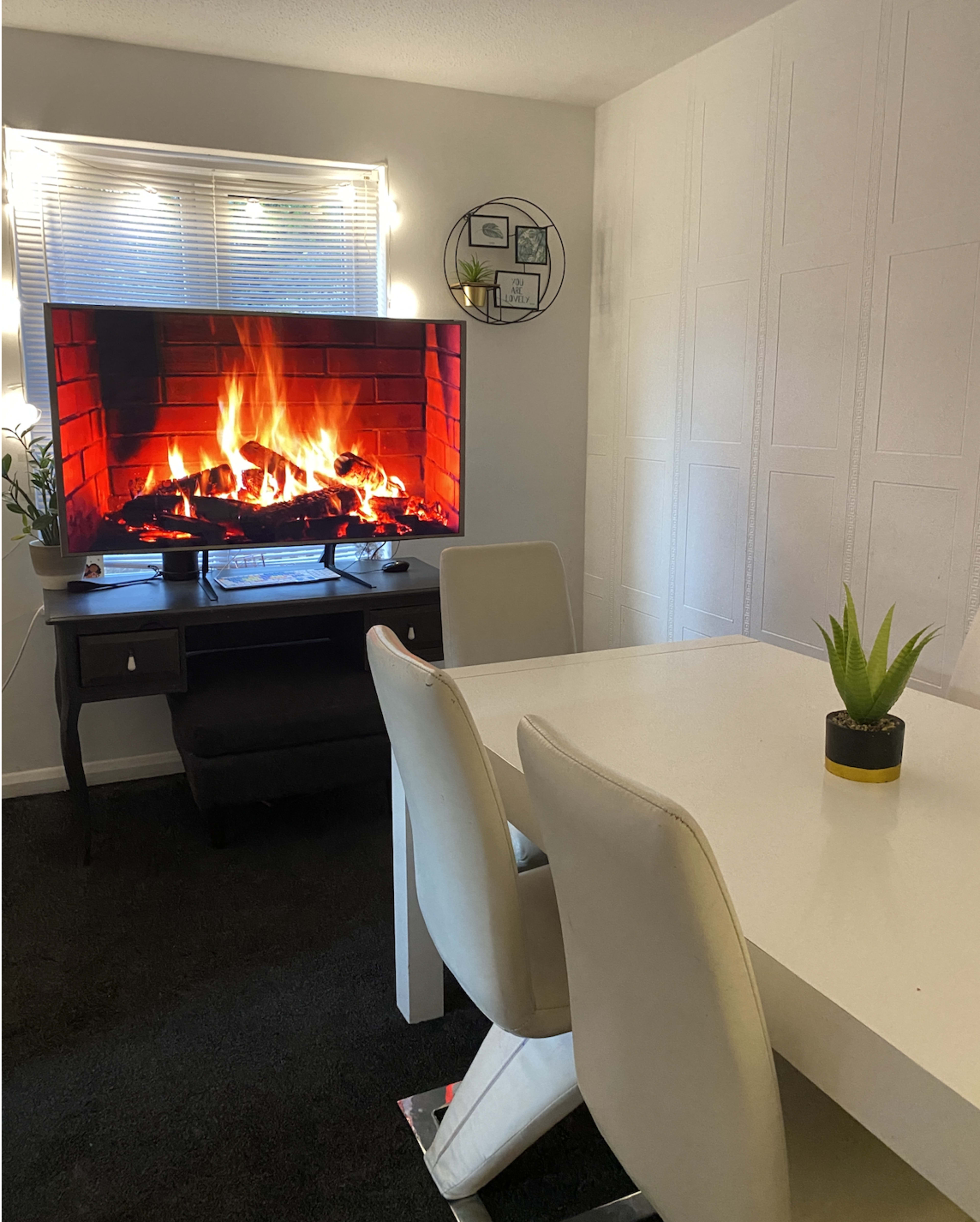 A television displaying a fireplace scene is positioned beside a white dining table and chairs in a well-lit room.
