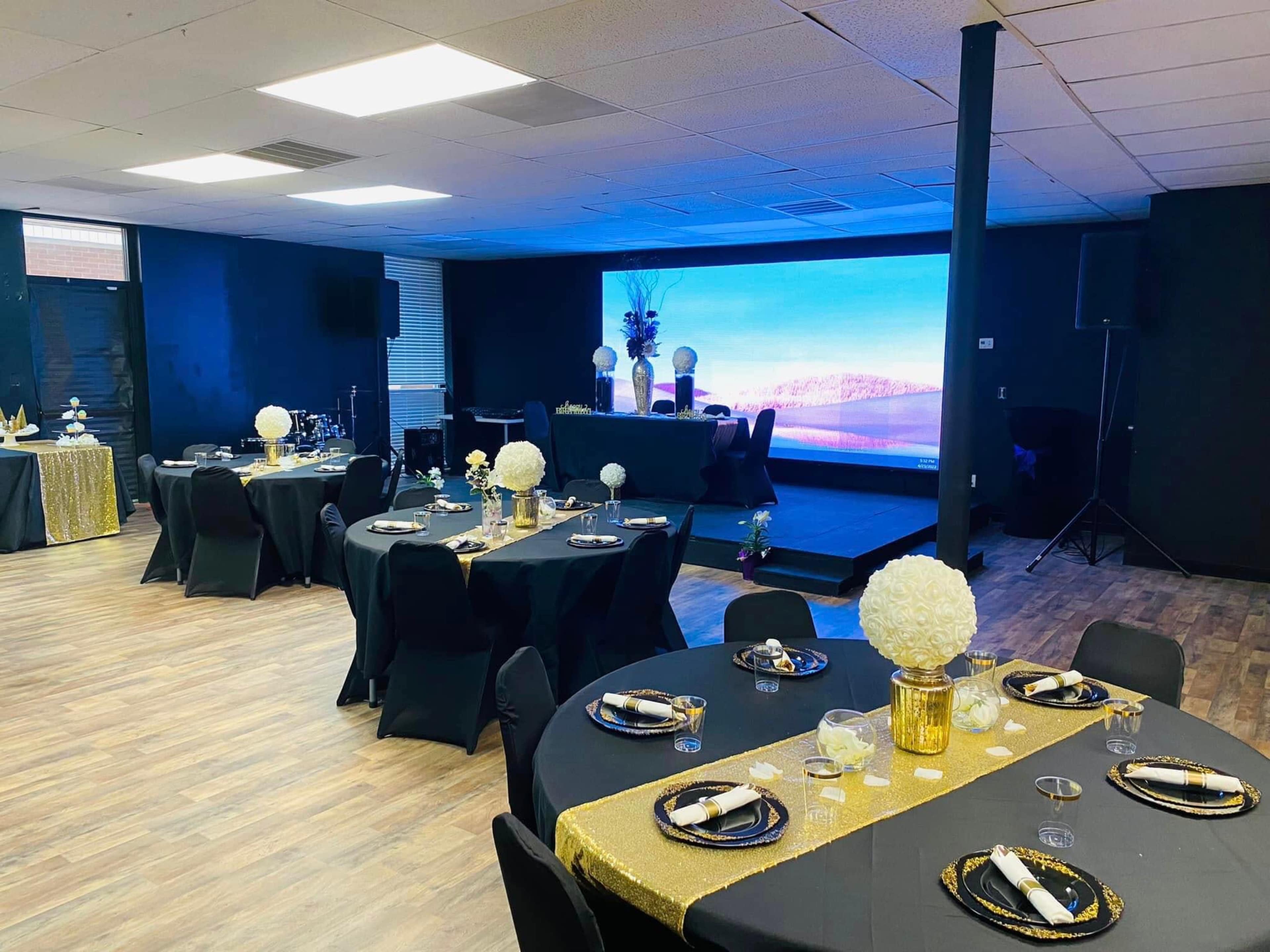 A banquet hall set up for an event, with black-tabled seating arrangements, decorative centerpieces, and a stage featuring a large screen in the background.