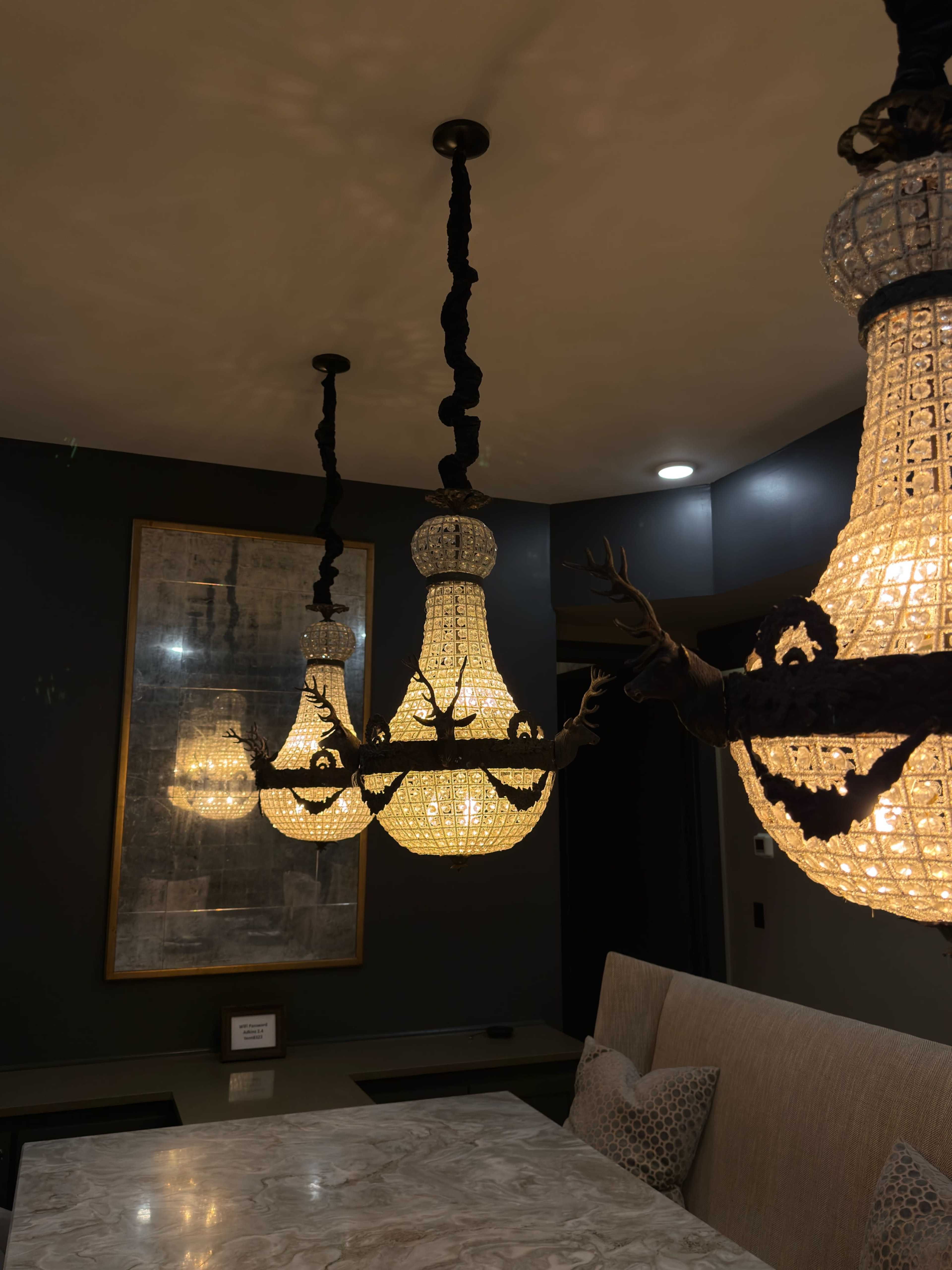 The image shows three ornate chandeliers hanging from the ceiling in a dimly lit dining area, with a patterned wall in the background and a marble table in the foreground.