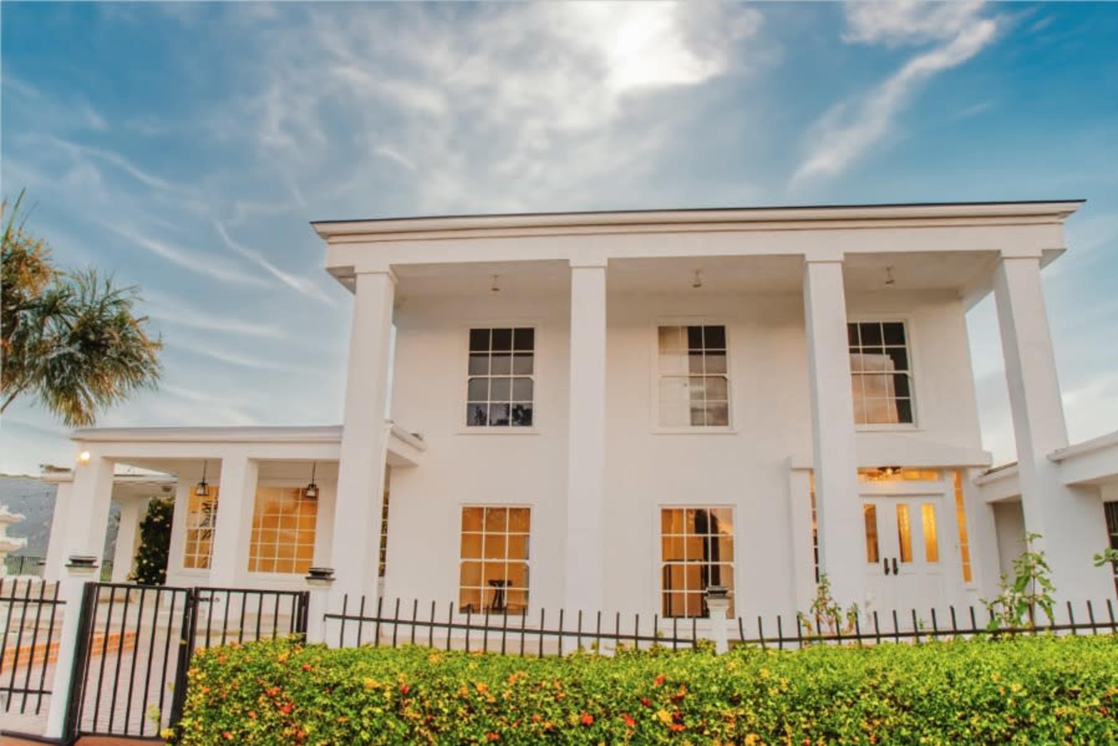 A two-story white house with tall columns and large windows, surrounded by a black metal fence and landscaped with green plants.