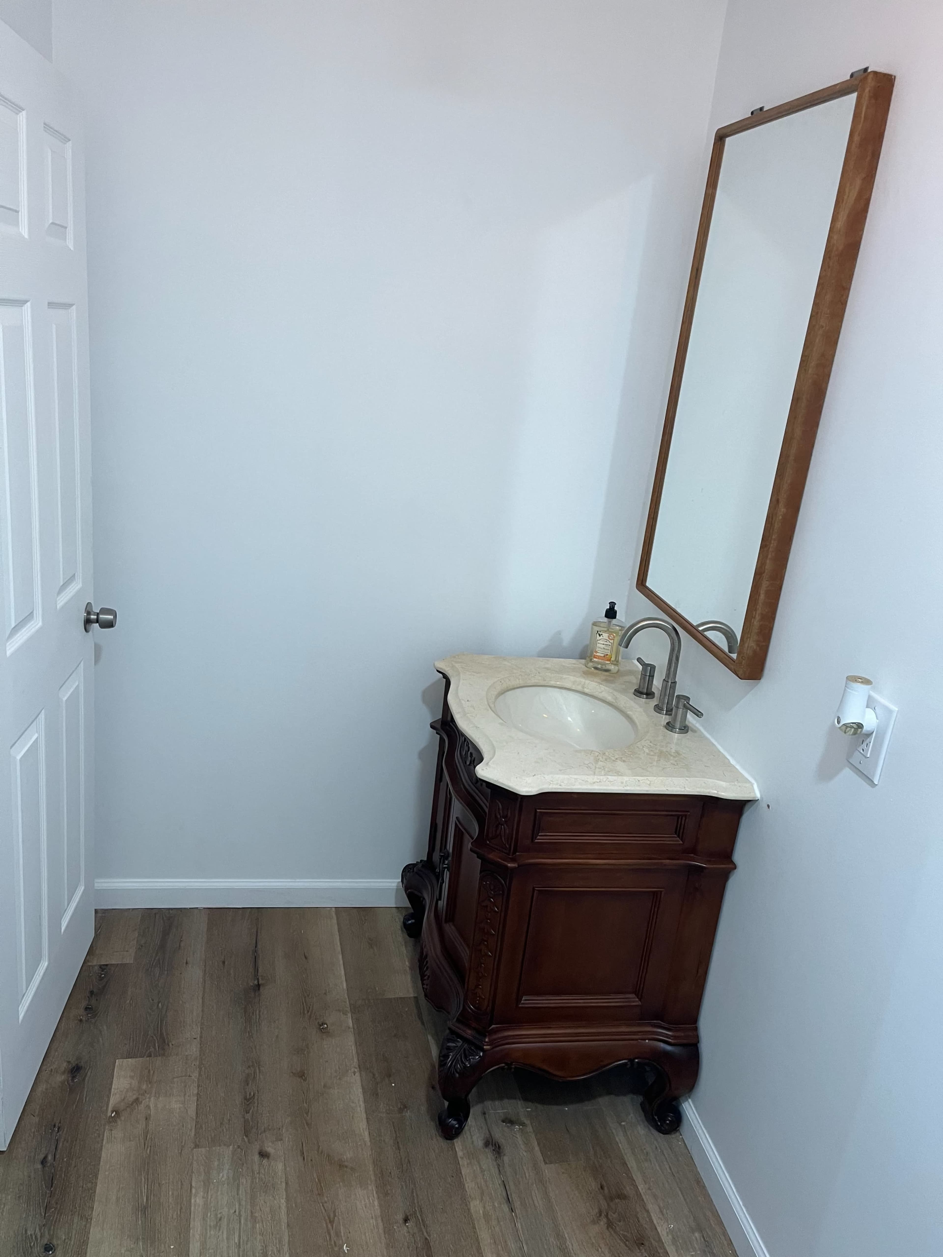 A bathroom featuring a wooden vanity with a marble sink and a large mirror above it, set against plain white walls and a wooden floor.