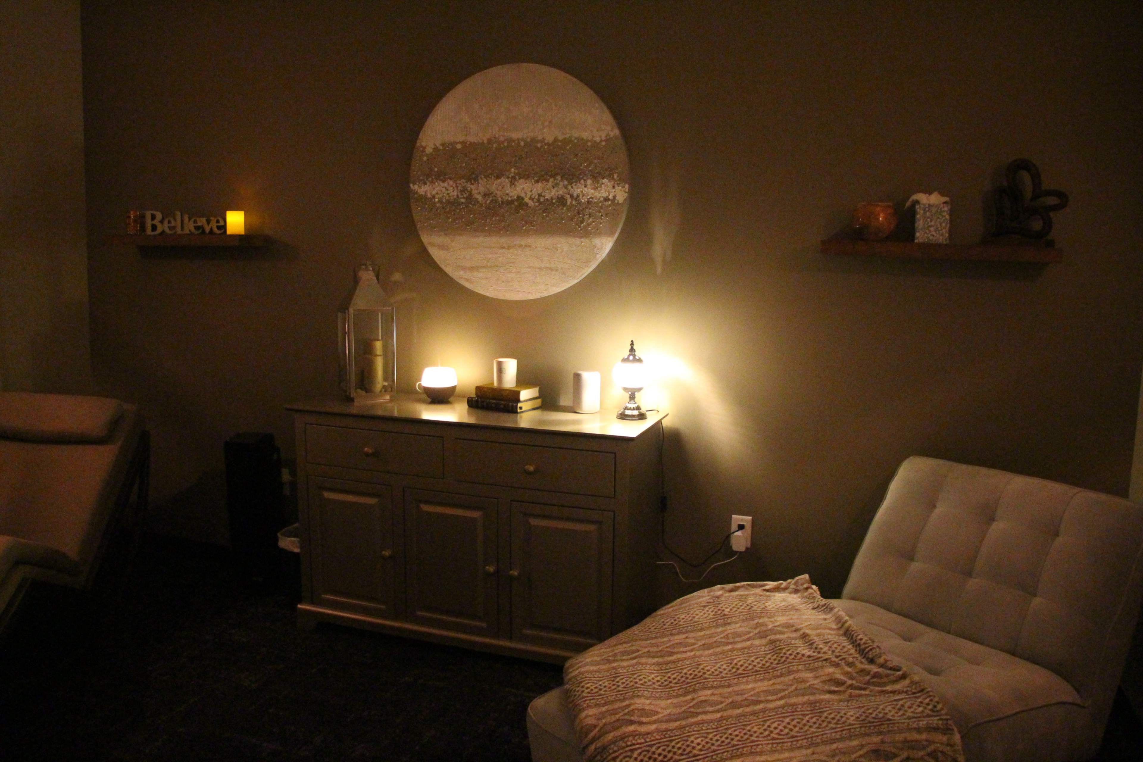 A cozy interior scene featuring a light gray chest of drawers beside an armchair, with a round wall art piece, candles, and decorative items on shelves.